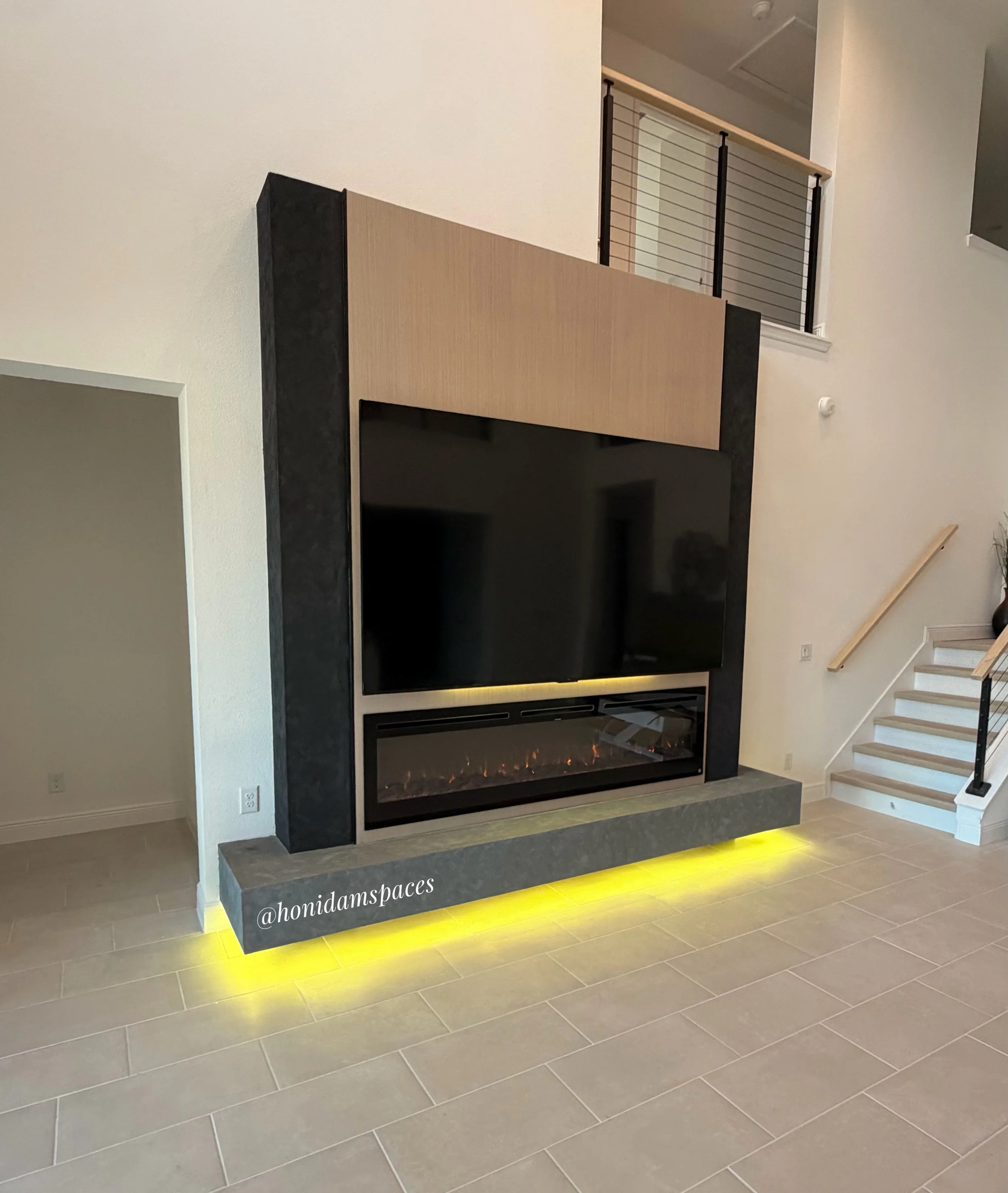 Modern indoor fireplace with a large black frame, beige wooden panel, and yellow lighting underneath, located in a room with gray tiled flooring and white walls.