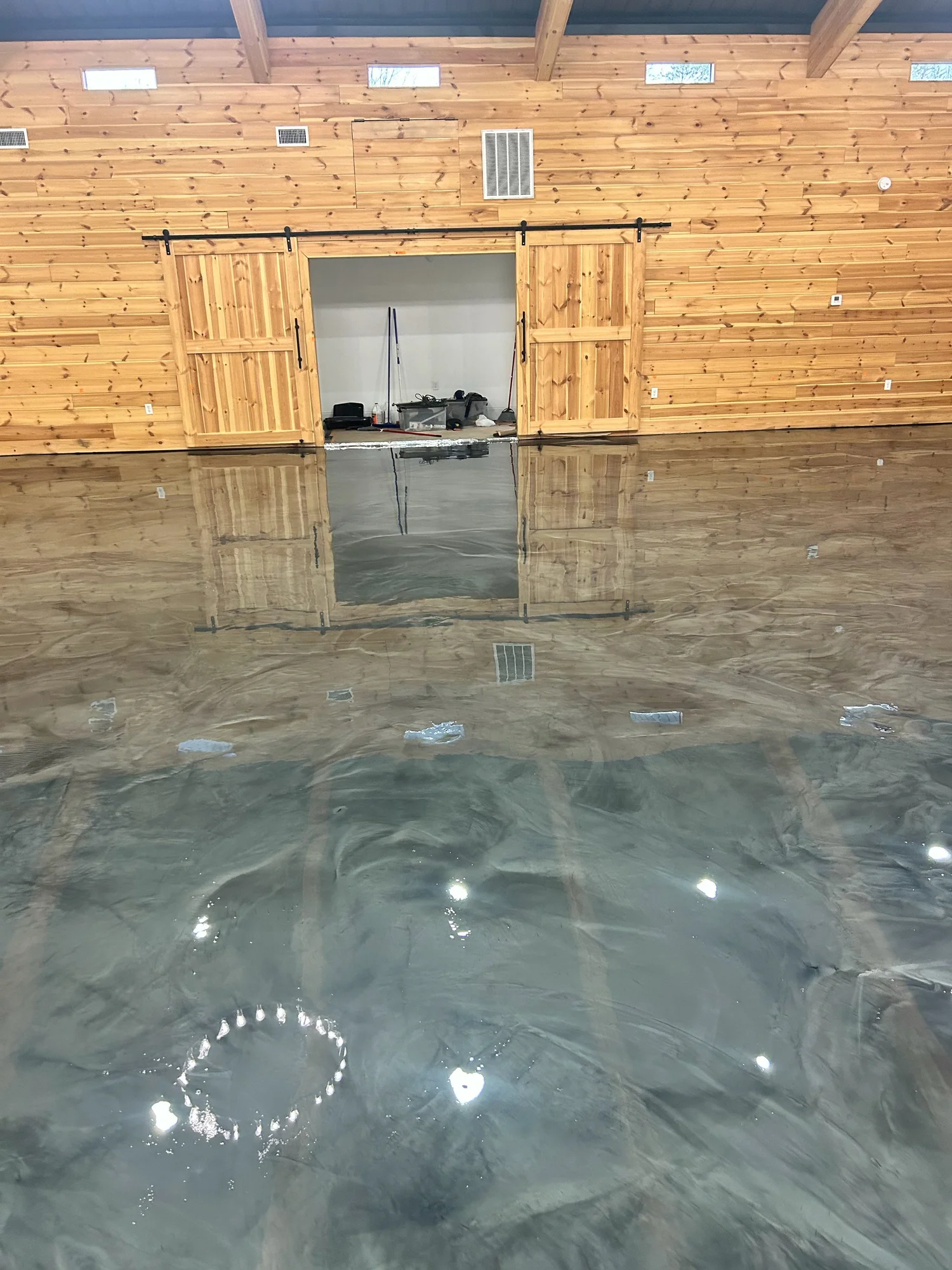 Indoor wooden building with a large, polished concrete floor, sliding barn doors open to a storage room with cleaning supplies, and high windows letting in natural light.