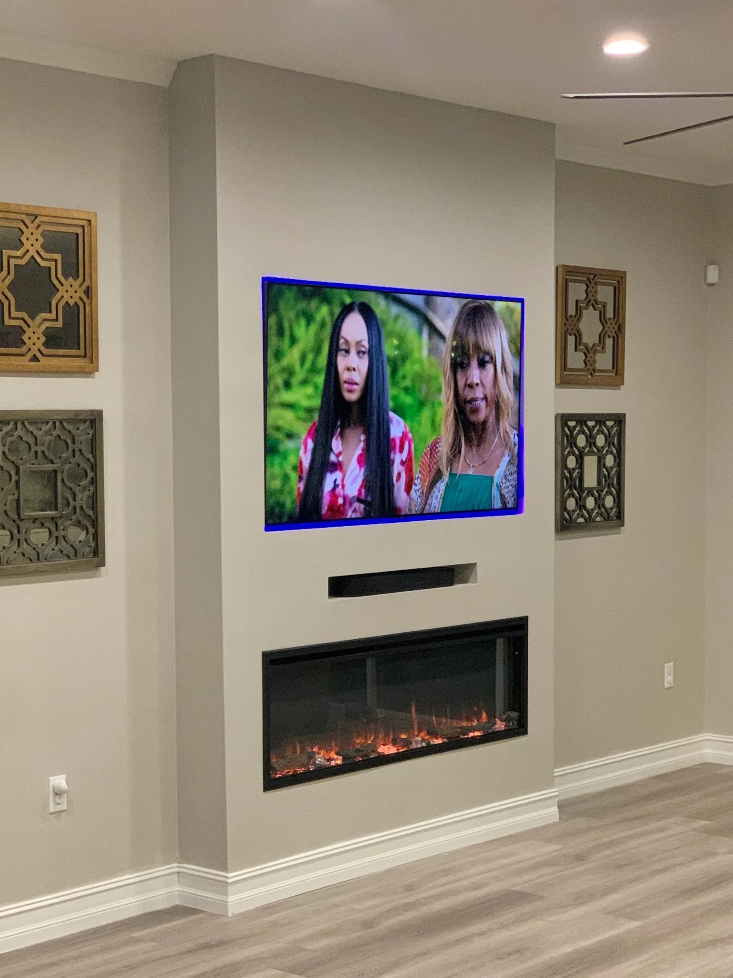 Living room with a wall-mounted television displaying two women, a built-in electric fireplace below the TV, and decorative wall art pieces on the walls.