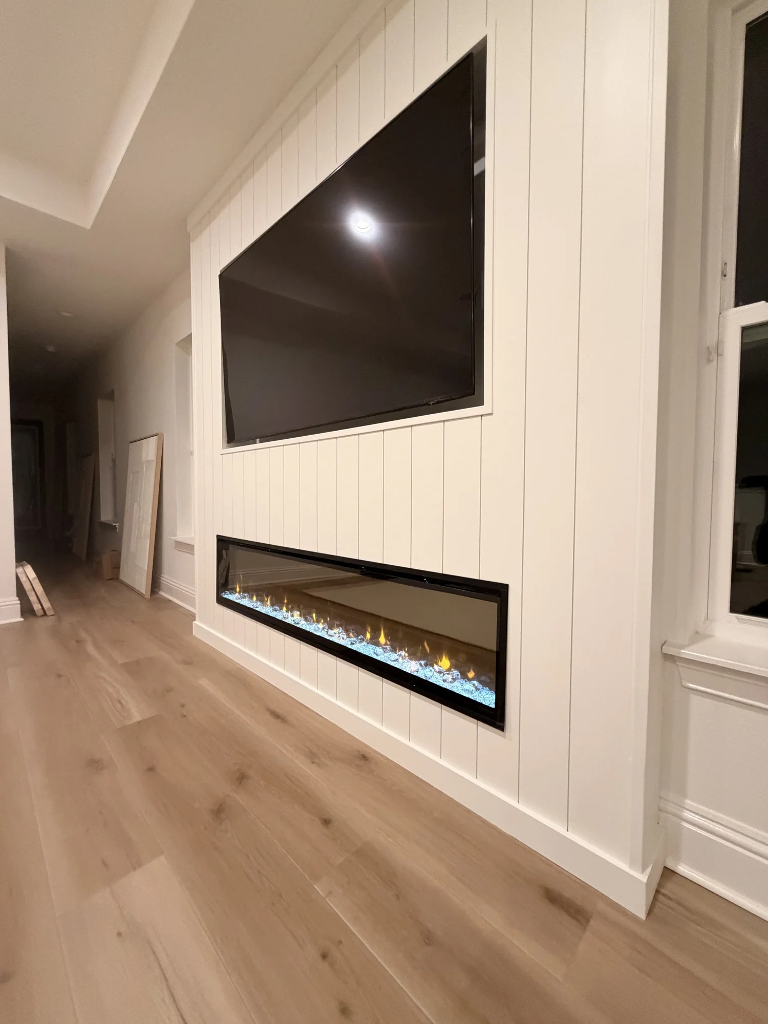 Living room with a wall-mounted TV above a built-in electric fireplace with decorative flames and blue accents. Light wood flooring and white paneled walls with a window on the right.