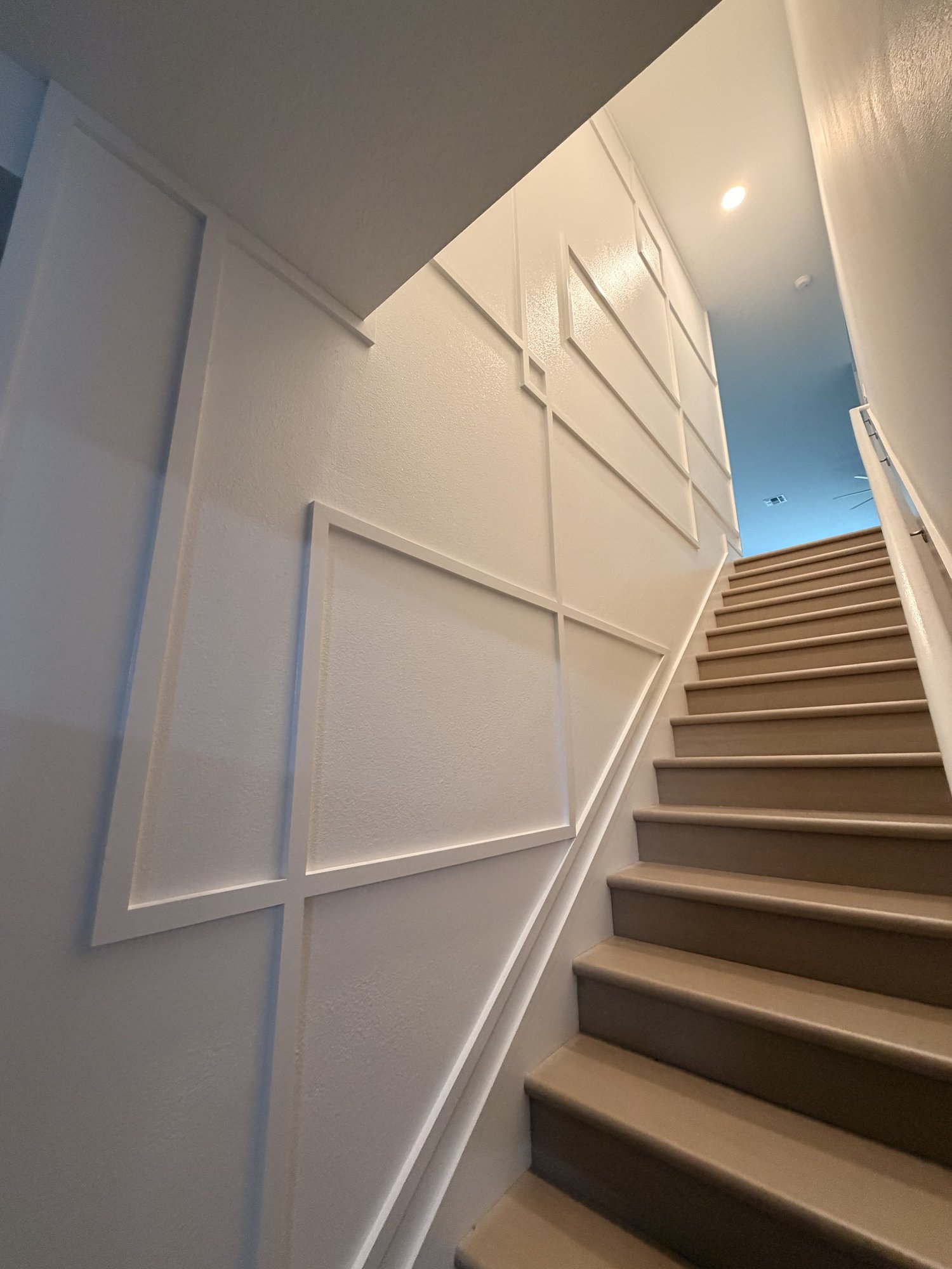Interior view of a staircase with wooden steps, a textured white wall decorated with rectangular panels, and a ceiling with recessed lighting.