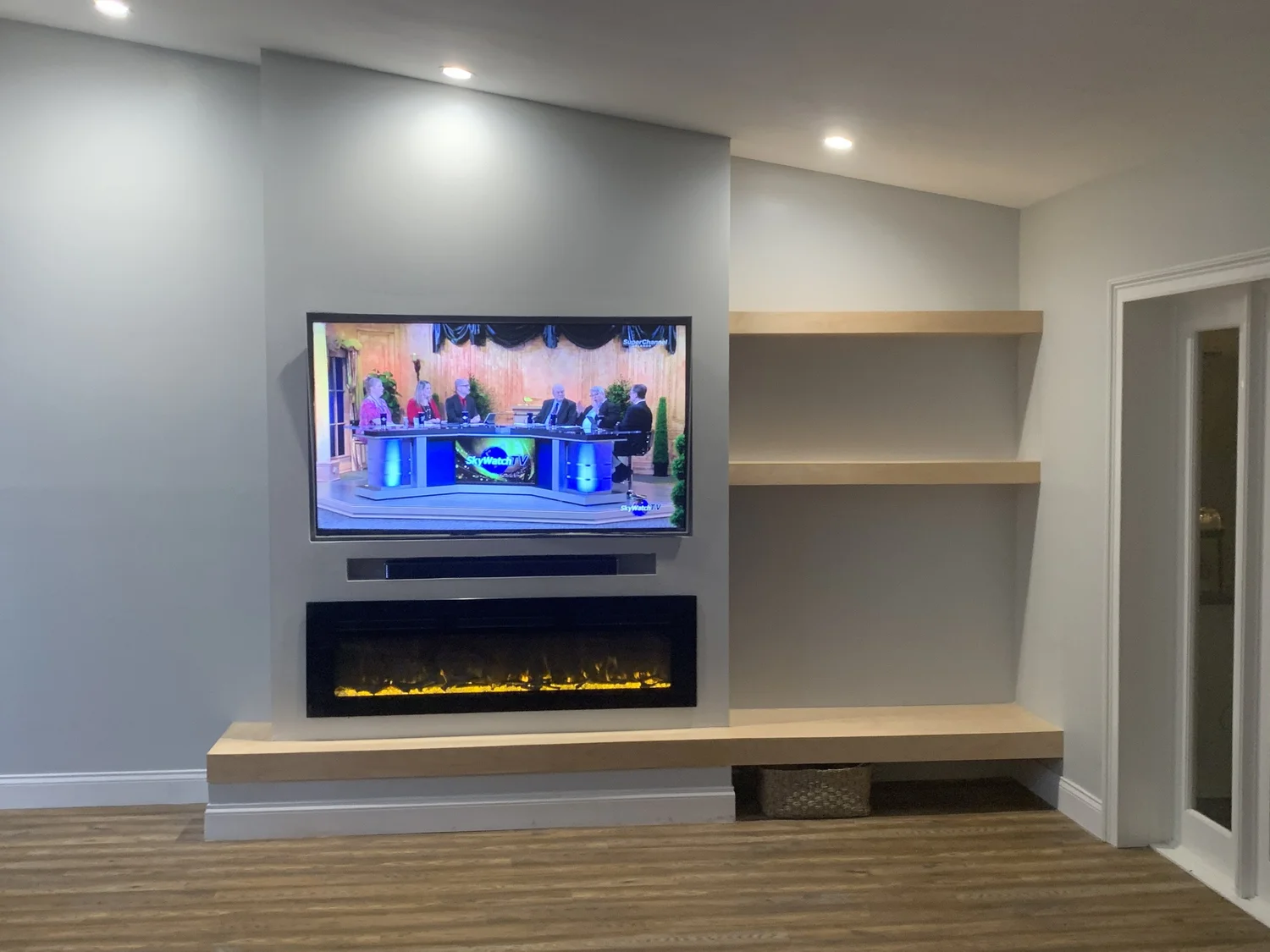 Living room with wall-mounted flat-screen TV displaying a panel discussion, modern electric fireplace below the TV, and empty built-in wooden shelves to the right, with a doorway visible on the far right.