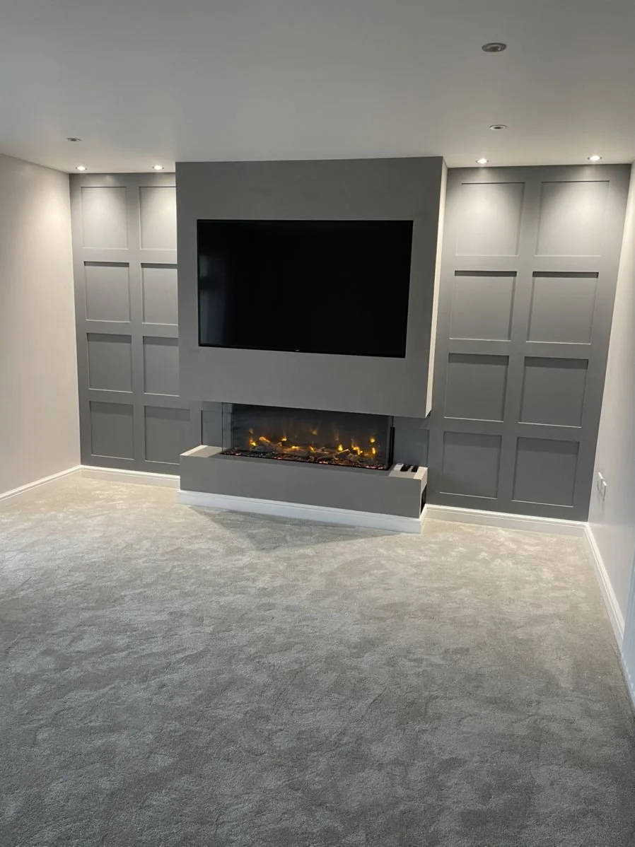 Modern living room with a gray wall, mounted flat-screen TV, and a built-in electric fireplace below the TV. The room has recessed ceiling lights and light gray carpet flooring.