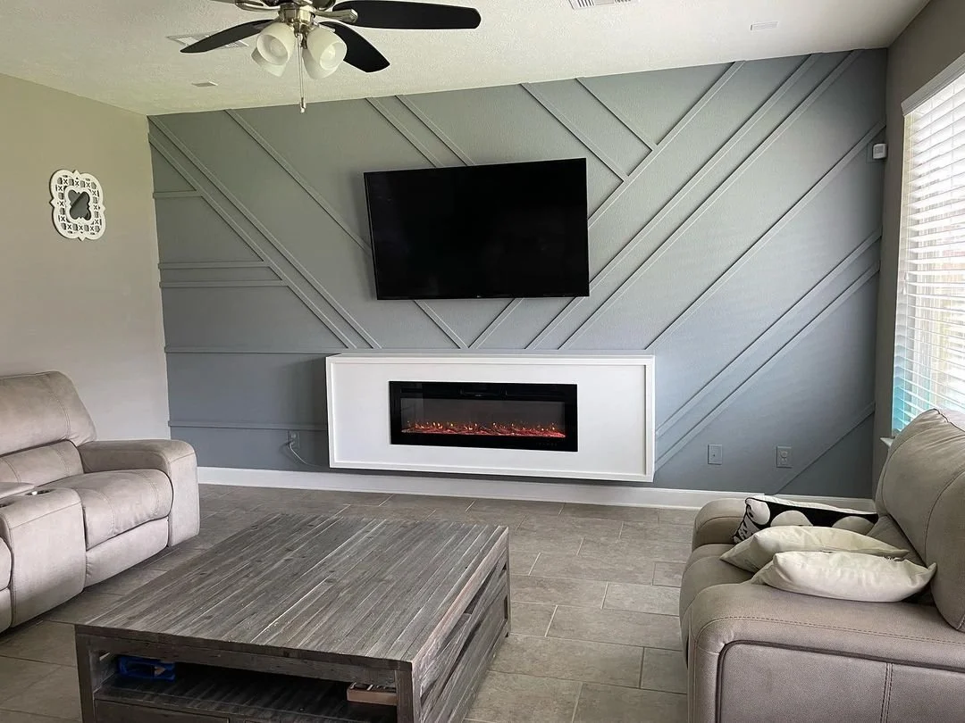 Living room with a gray accent wall, a flat-screen TV mounted above an electric fireplace, two beige sofas, a wooden coffee table, ceiling fan, and a window with blinds.