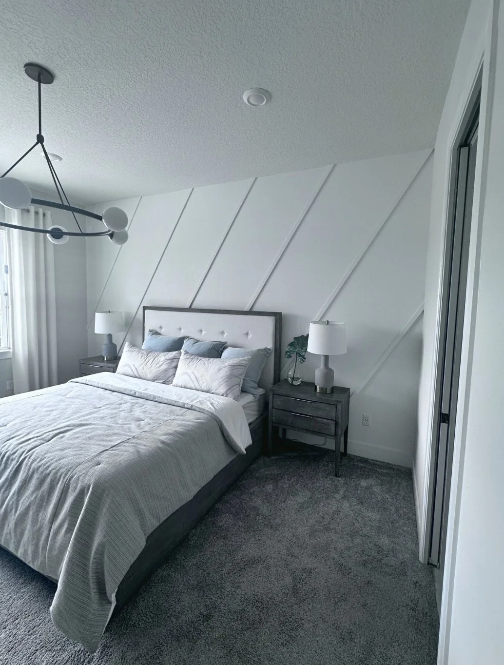 A modern bedroom with a bed, nightstand, and lamp, situated under a sloped wall with wooden paneling, and a contemporary chandelier hanging from the ceiling.
