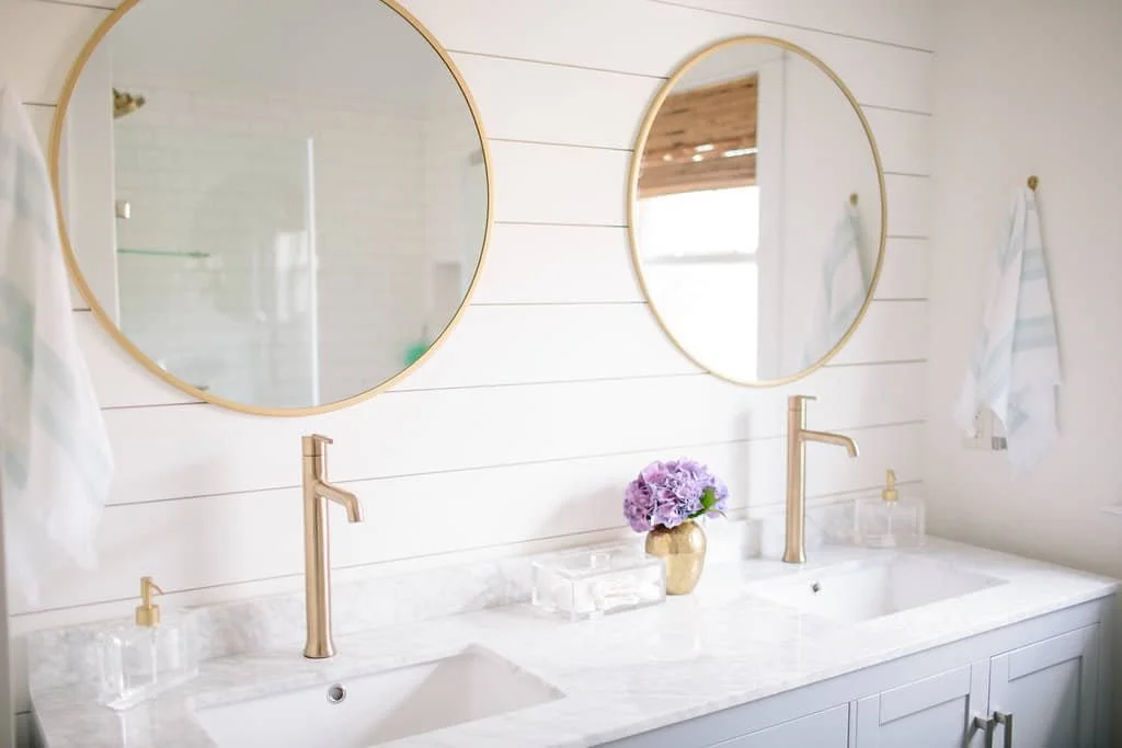 Bathroom with two round gold-framed mirrors above a white marble double sink vanity, decorated with a purple hydrangea flower in a gold vase, white towel hooks with white towels, and clear soap dispensers.