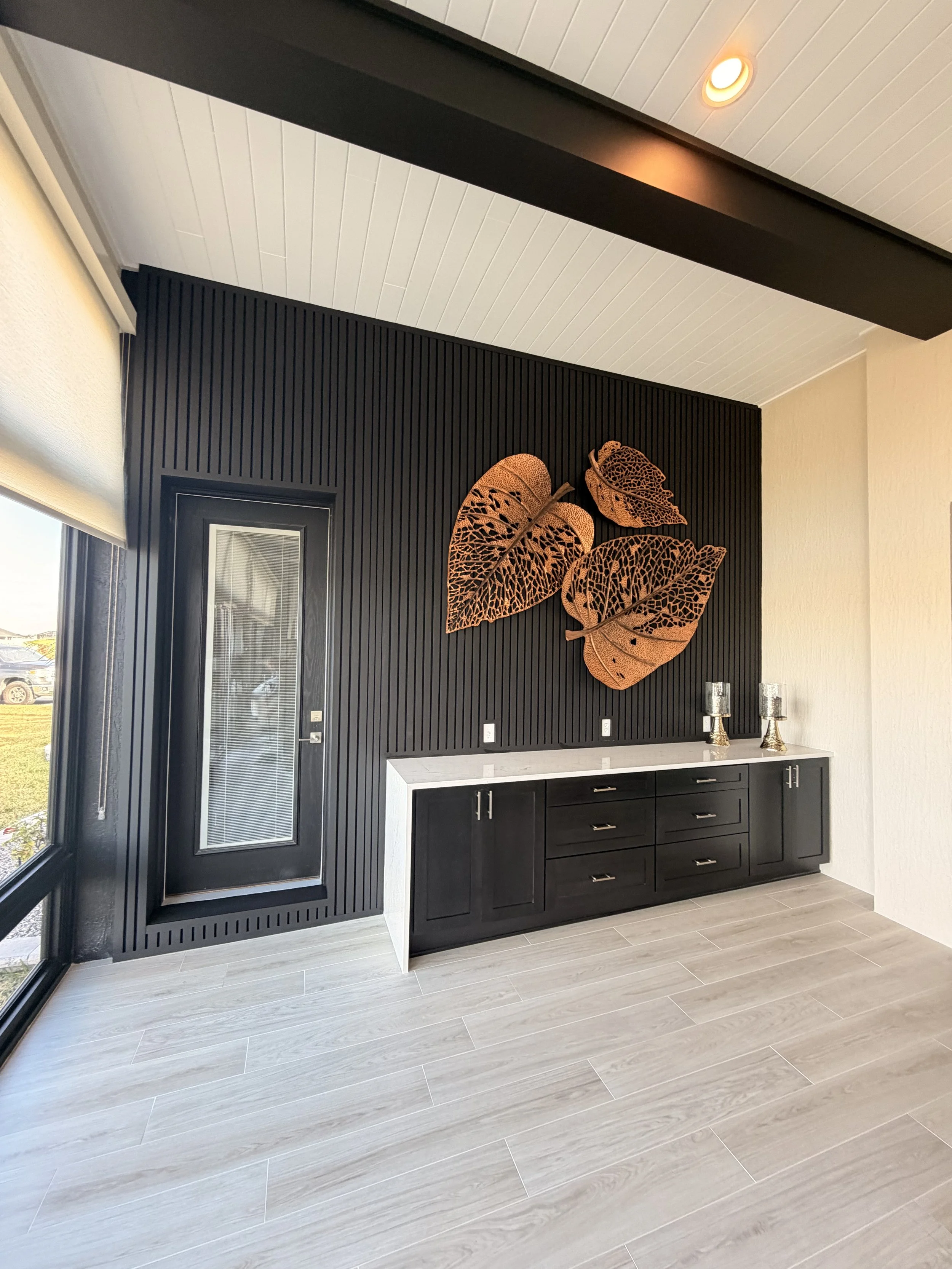 Modern interior room featuring a black accent wall with gold decorative leaves, a white marble countertop, black cabinets, two decorative vases, and large windows with natural light.