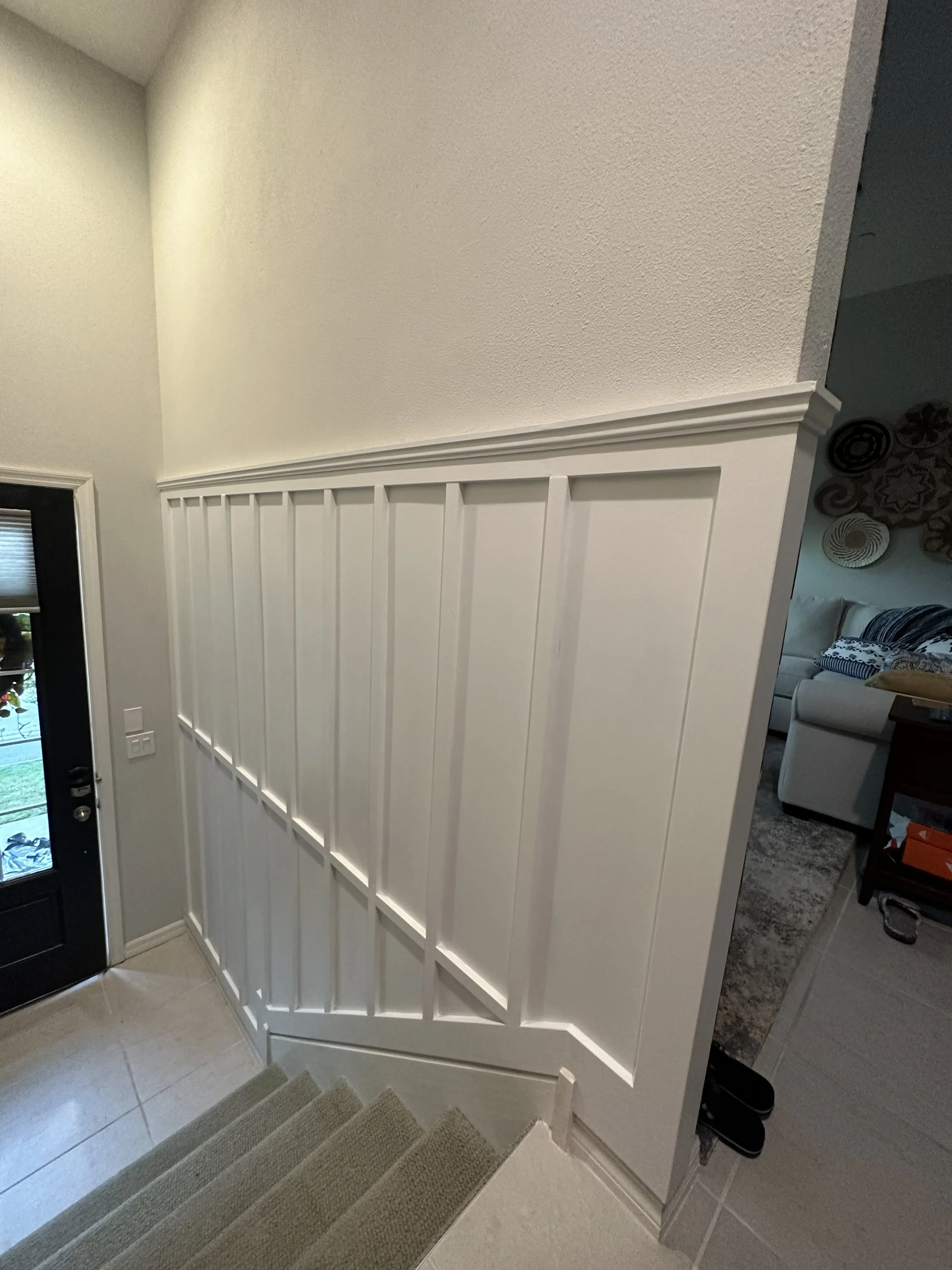 Interior view of a staircase landing with white wooden wainscoting, beige carpeted stairs, and a black door with a window to the left. Part of a living room with sectional sofa and decor is visible in the background.