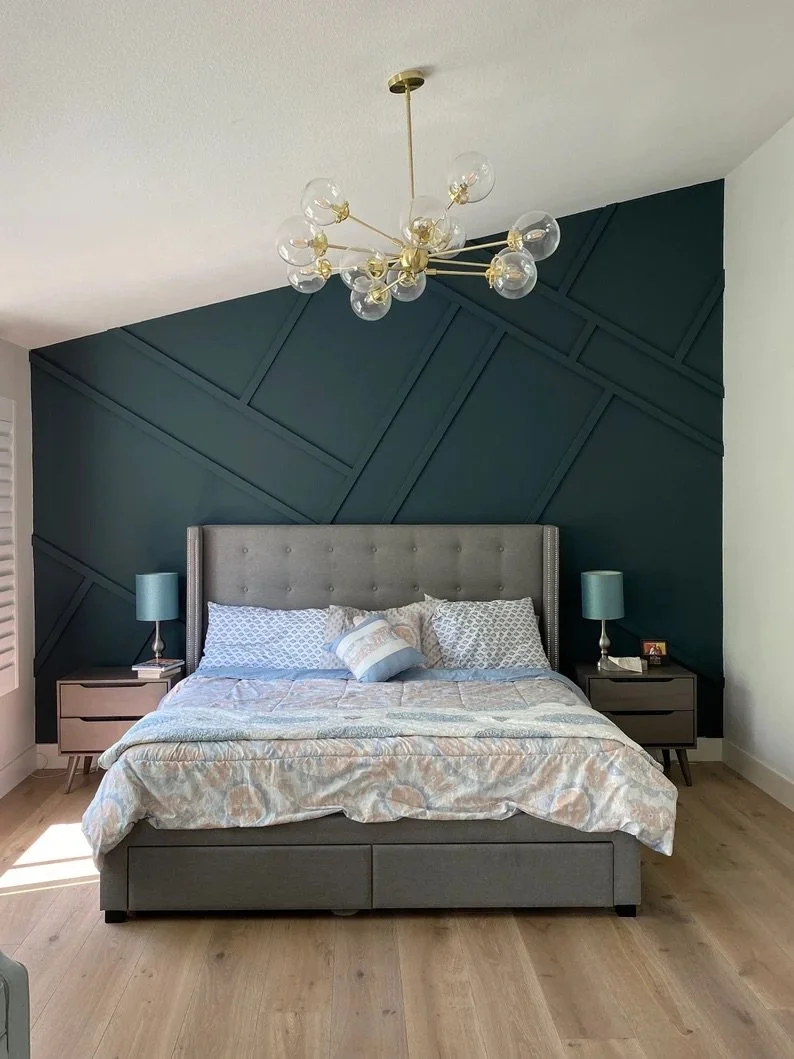 A modern bedroom with a gray upholstered bed, blue pillow cushions, and a buff-colored bedspread. Two matching nightstands with blue lamps, and an abstract geometric dark green accent wall behind the bed. A gold and glass chandelier hangs from the ce