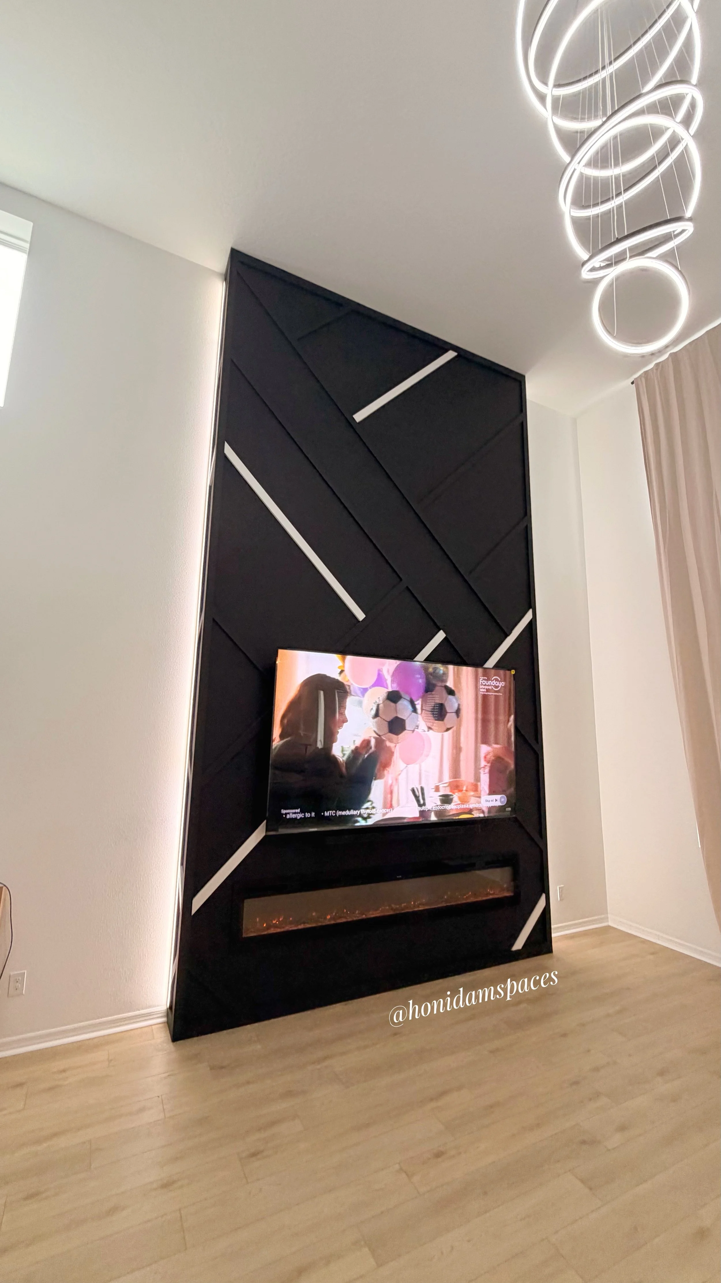 Living room with a black geometric patterned wall behind a TV and fireplace, a modern circular chandelier on the ceiling, a window with curtains, and light wood floor.