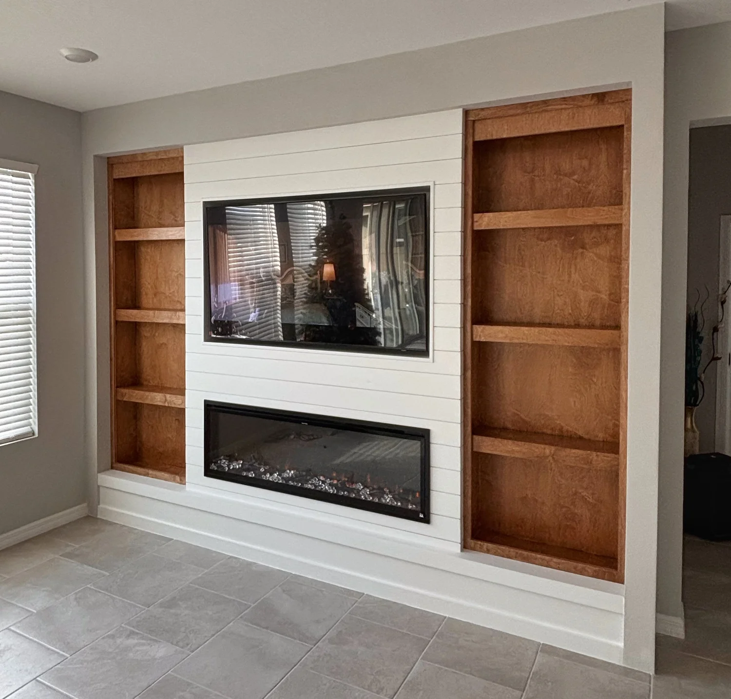 Living room with built-in wooden bookshelves and a modern electric fireplace, with beige tiled flooring and a window with blinds on the left side.