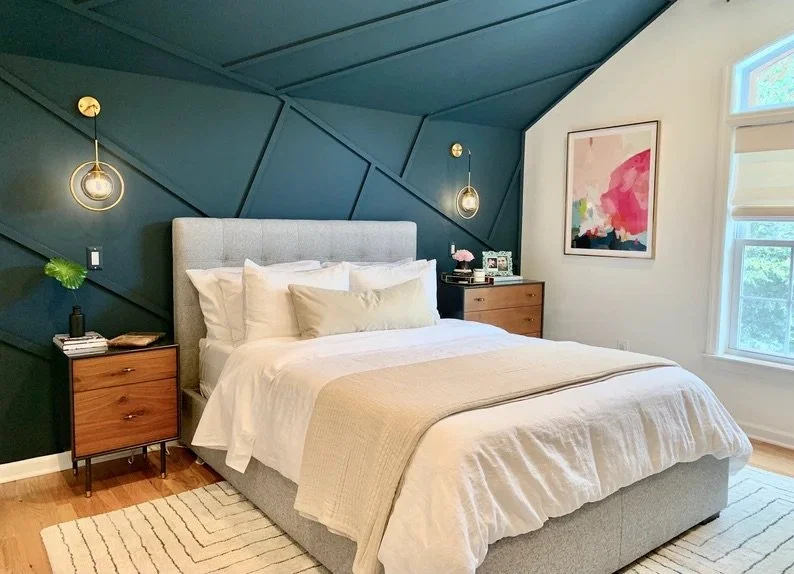 Bedroom with a gray upholstered bed, white bedding, two nightstands with lamps and decor, dark blue geometric accent wall, colorful abstract art, and large window with natural light.