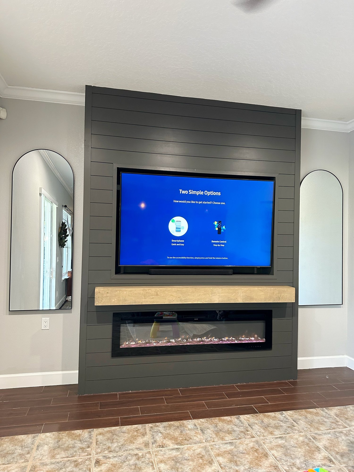 A living room corner featuring a wall-mounted TV displaying two options, with a modern electric fireplace below, framed by dark gray wooden paneling, and two tall, arched mirrors on either side of the paneling.