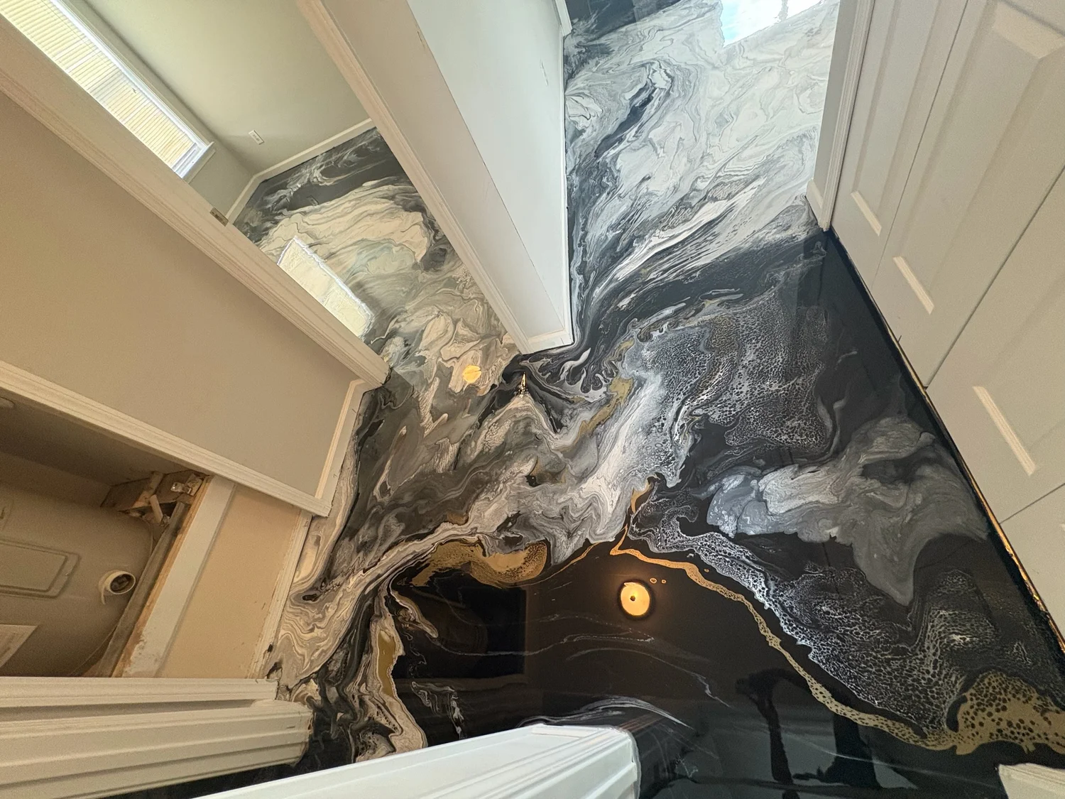 A room with black, white, gray, and gold marbled flooring and a wall mirror reflecting a window with blinds.