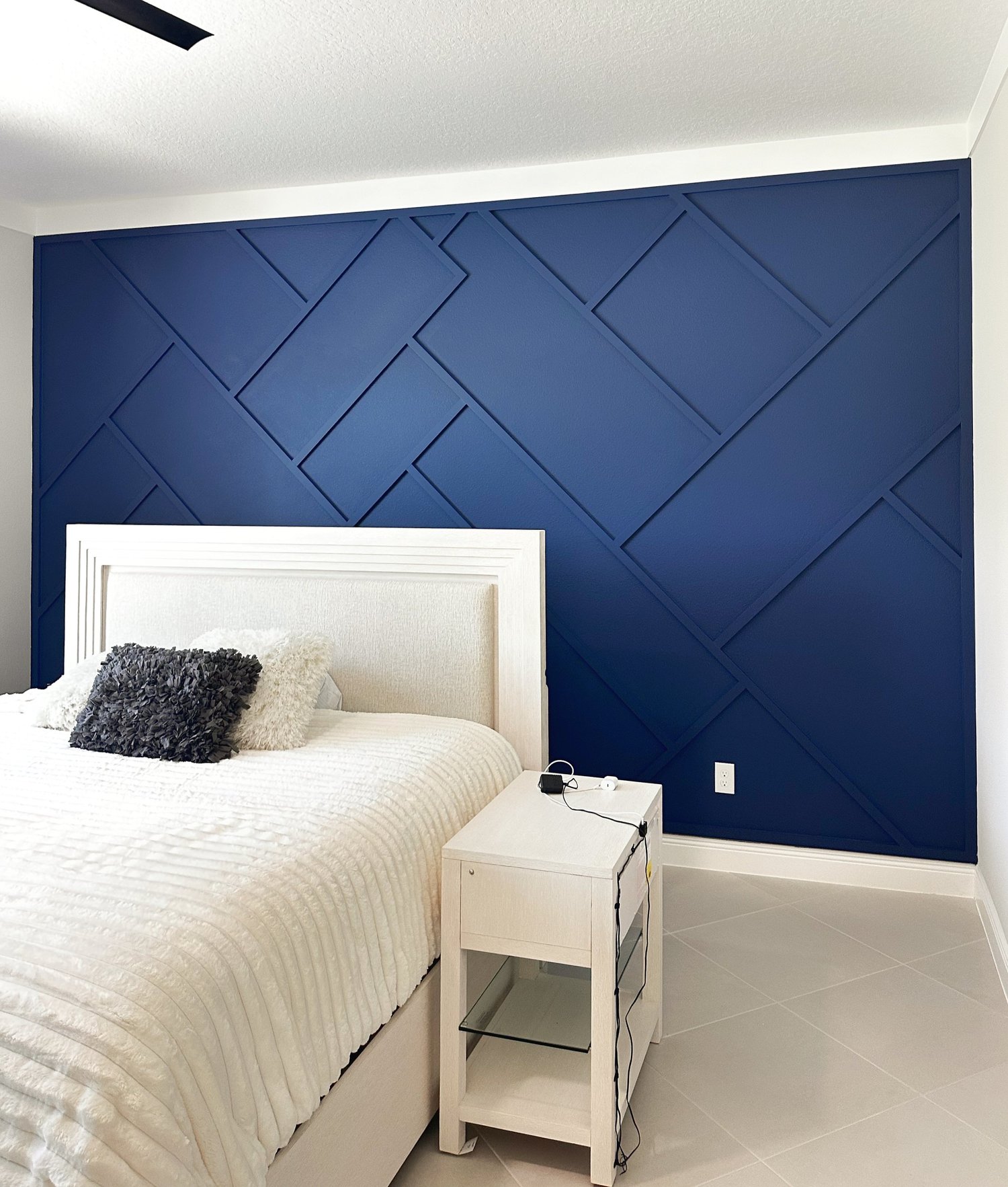 A bedroom with a blue accent wall featuring a geometric pattern, a white bed with a textured beige bedspread, and three decorative pillows, one black, one white, and one light green. There is a white nightstand with a black charger and a white power 