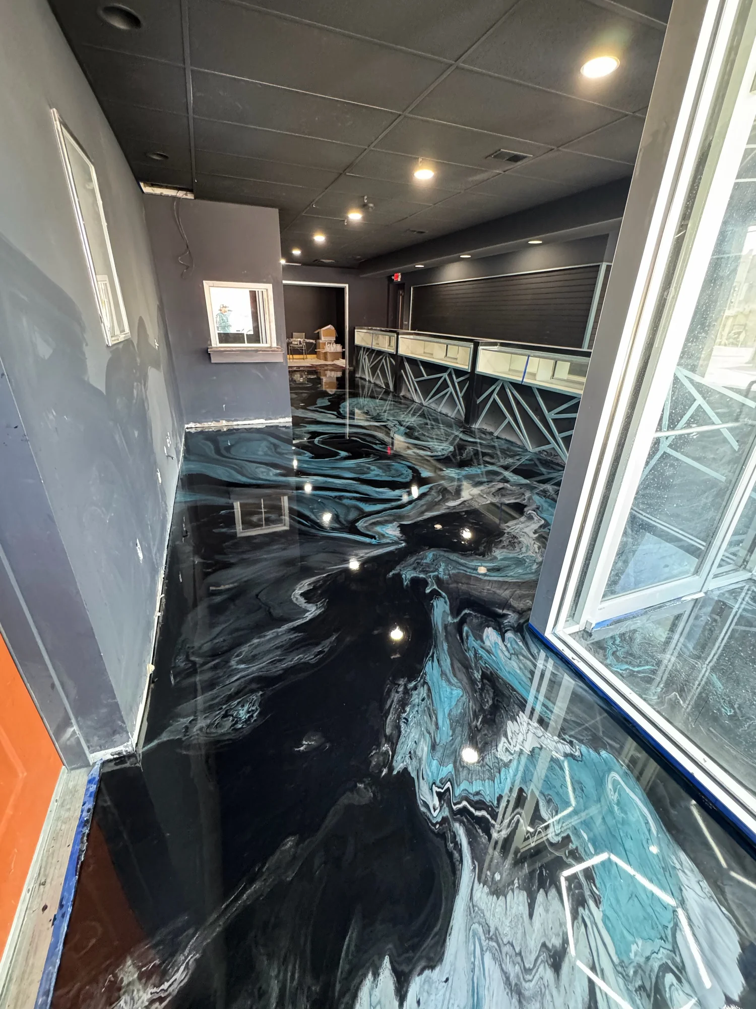 Interior of a building with a black marble floor, dark walls, ceiling lights, and a service counter with empty display cases, seen through a glass door.
