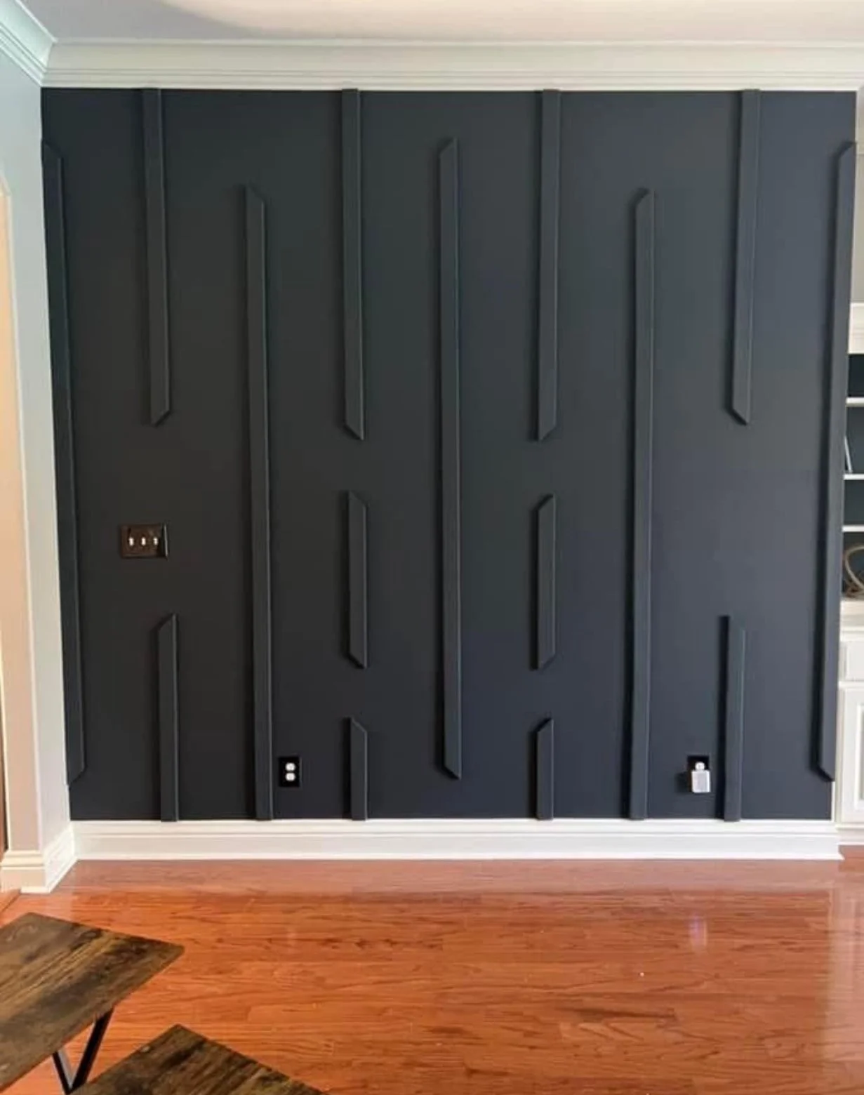 Interior wall painted dark, with decorative wooden paneling in raised geometric patterns. Electrical outlets along the wall and a white baseboard at the bottom. A section of hardwood floor and part of a wooden piece of furniture are visible in the fo