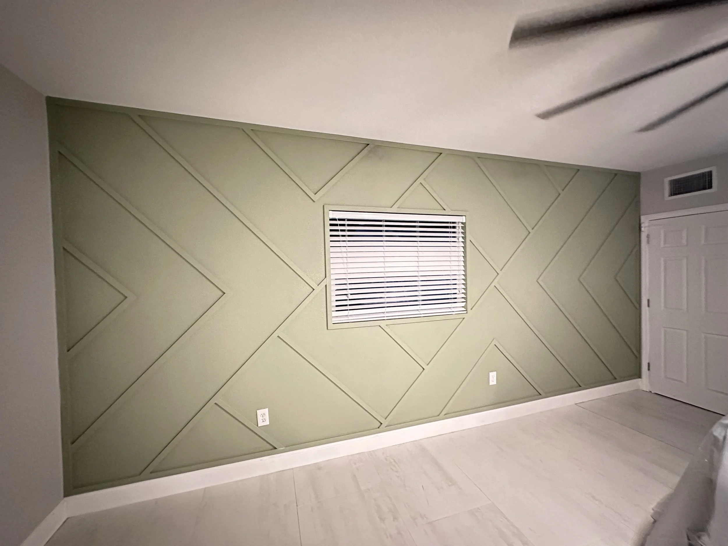 Empty room with green geometric accent wall, window with white blinds, white door, and white baseboards.