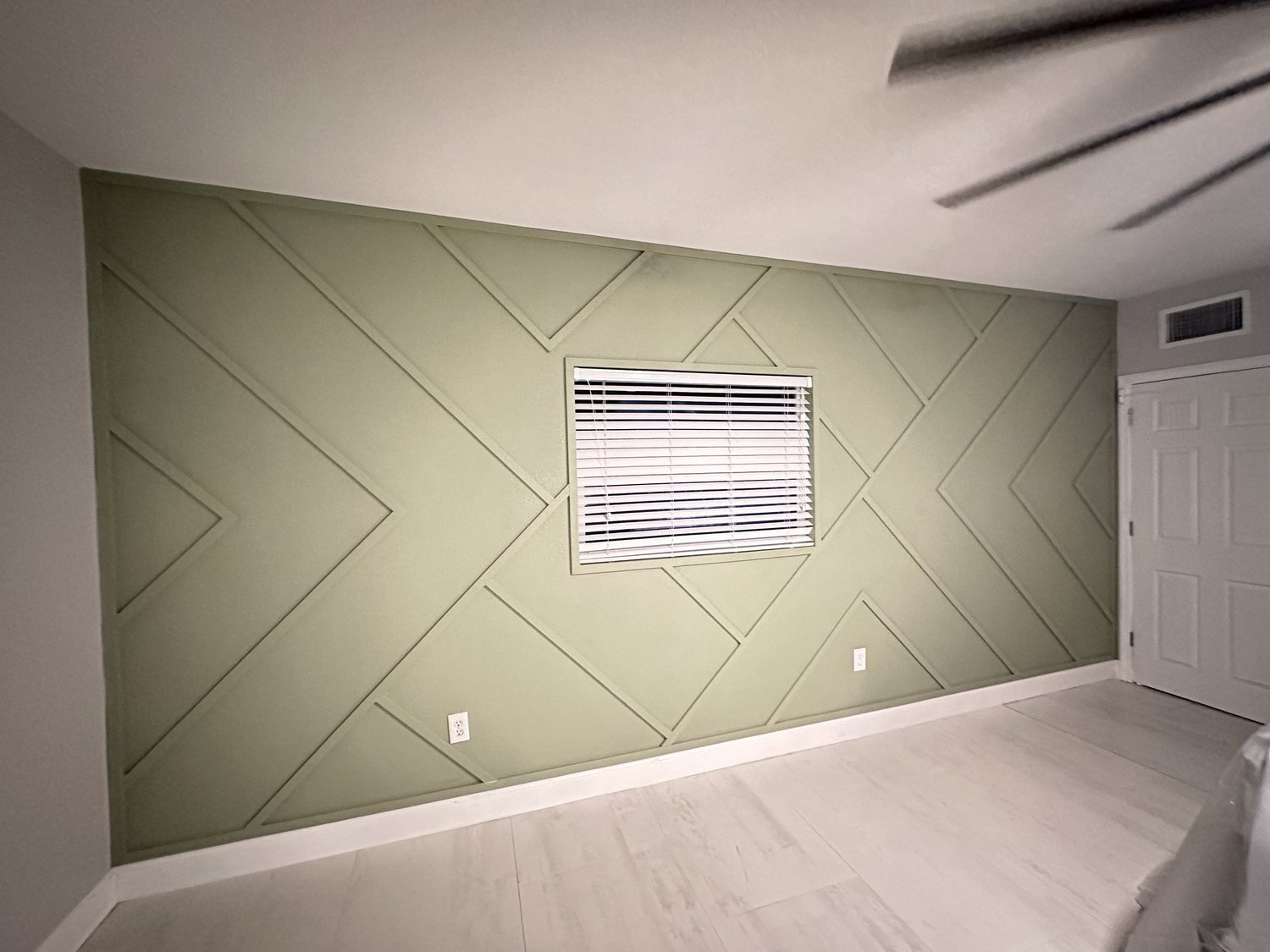 Empty room with green geometric accent wall, window with white blinds, white door, and white baseboards.