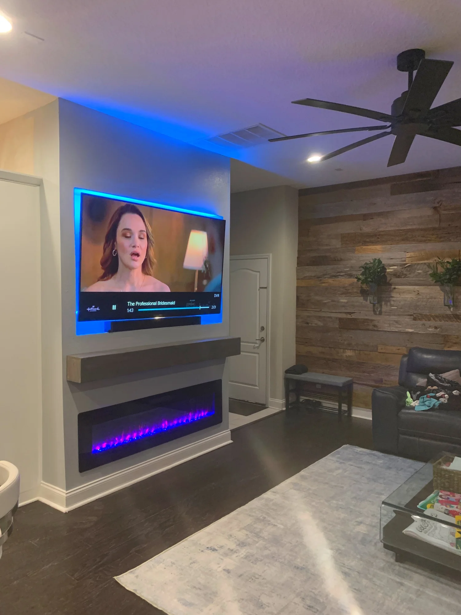 Living room with wall-mounted TV showing a woman, electric fireplace beneath, dark wood flooring, a light-colored rug, a black sofa with some items on it, a small table with items, and a wood-paneled accent wall with two hanging plants.