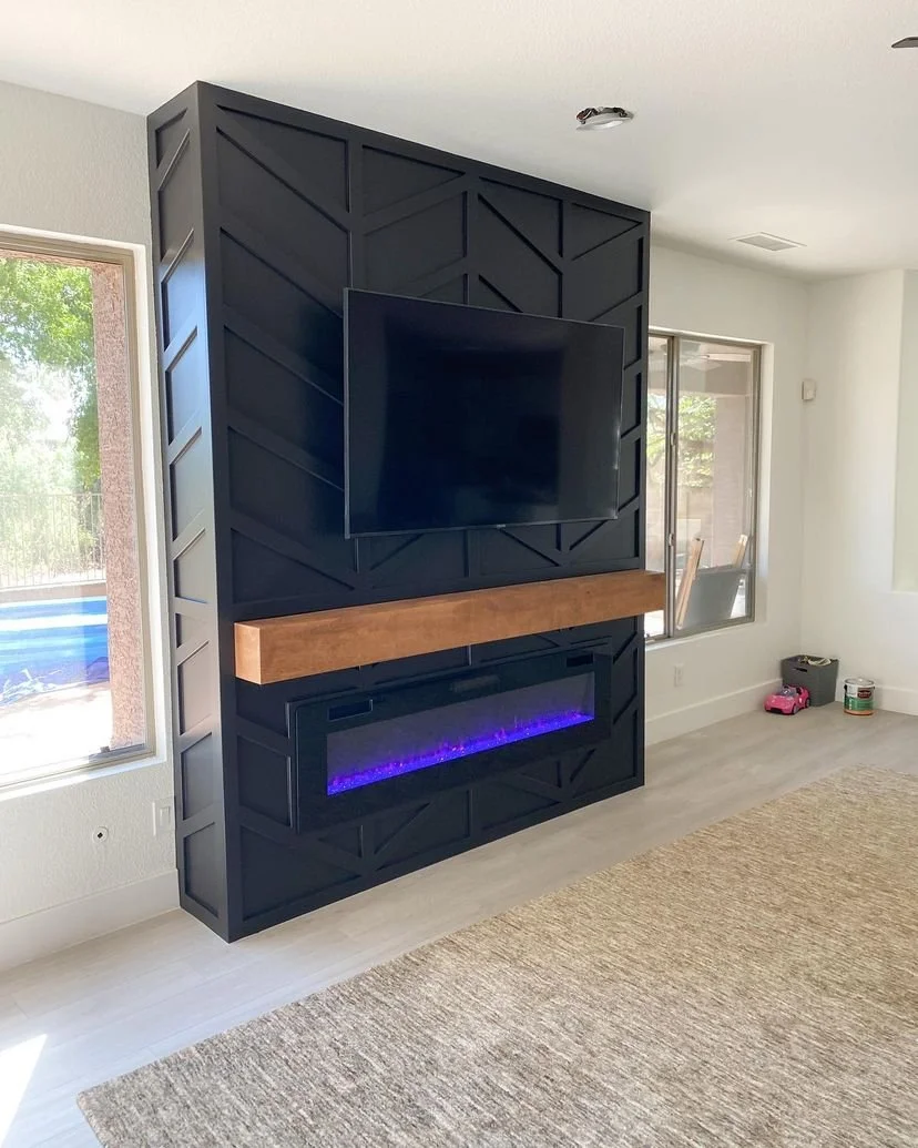 Modern living room with black wall featuring a mounted flat-screen TV and an electric fireplace with purple flames, accented with a wooden mantel, large windows, and a neutral-colored rug.