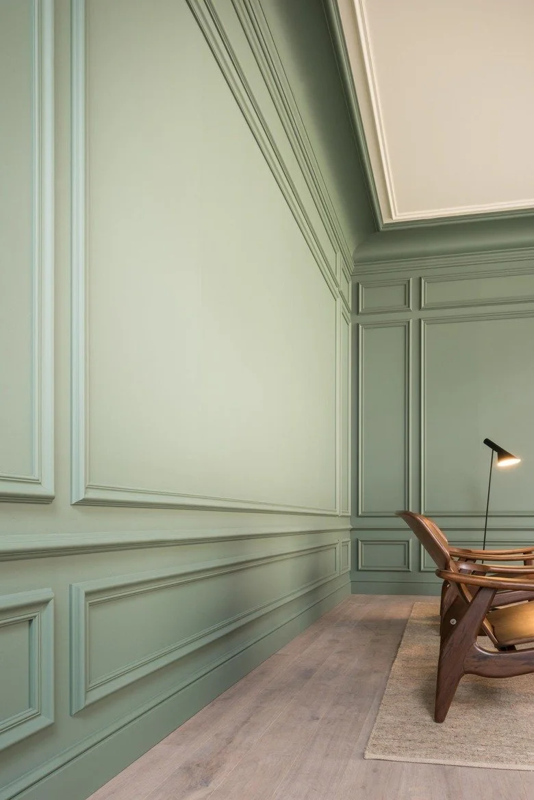 Interior corner of a room with green wainscoting and decorative molding, a wooden chair, a floor lamp, and a beige rug.