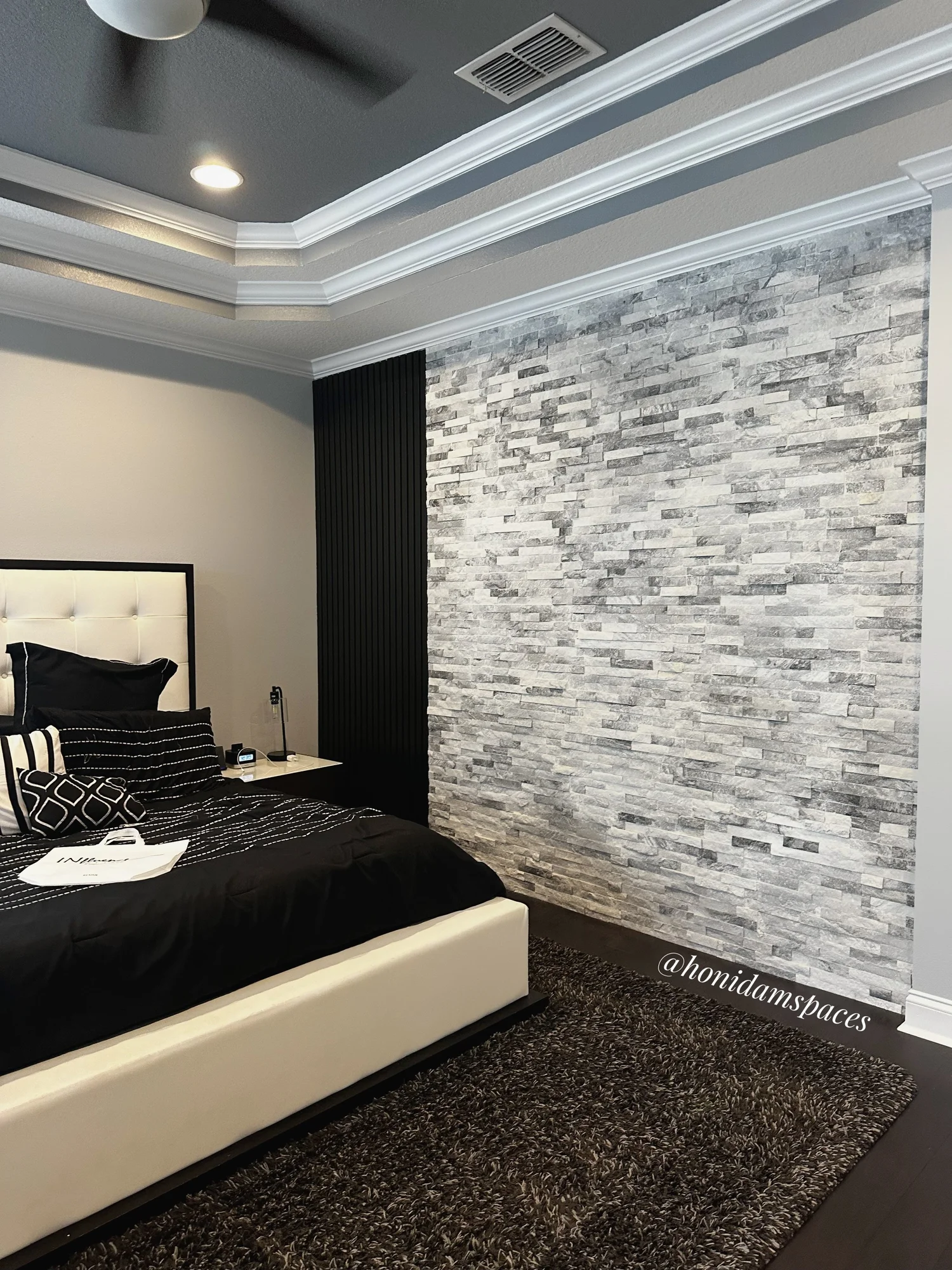 A modern bedroom features a white bed with black and white bedding, a textured gray stone wall, a black vertical panel, and a dark shaggy rug, with a bedside table containing a digital clock and a lamp.
