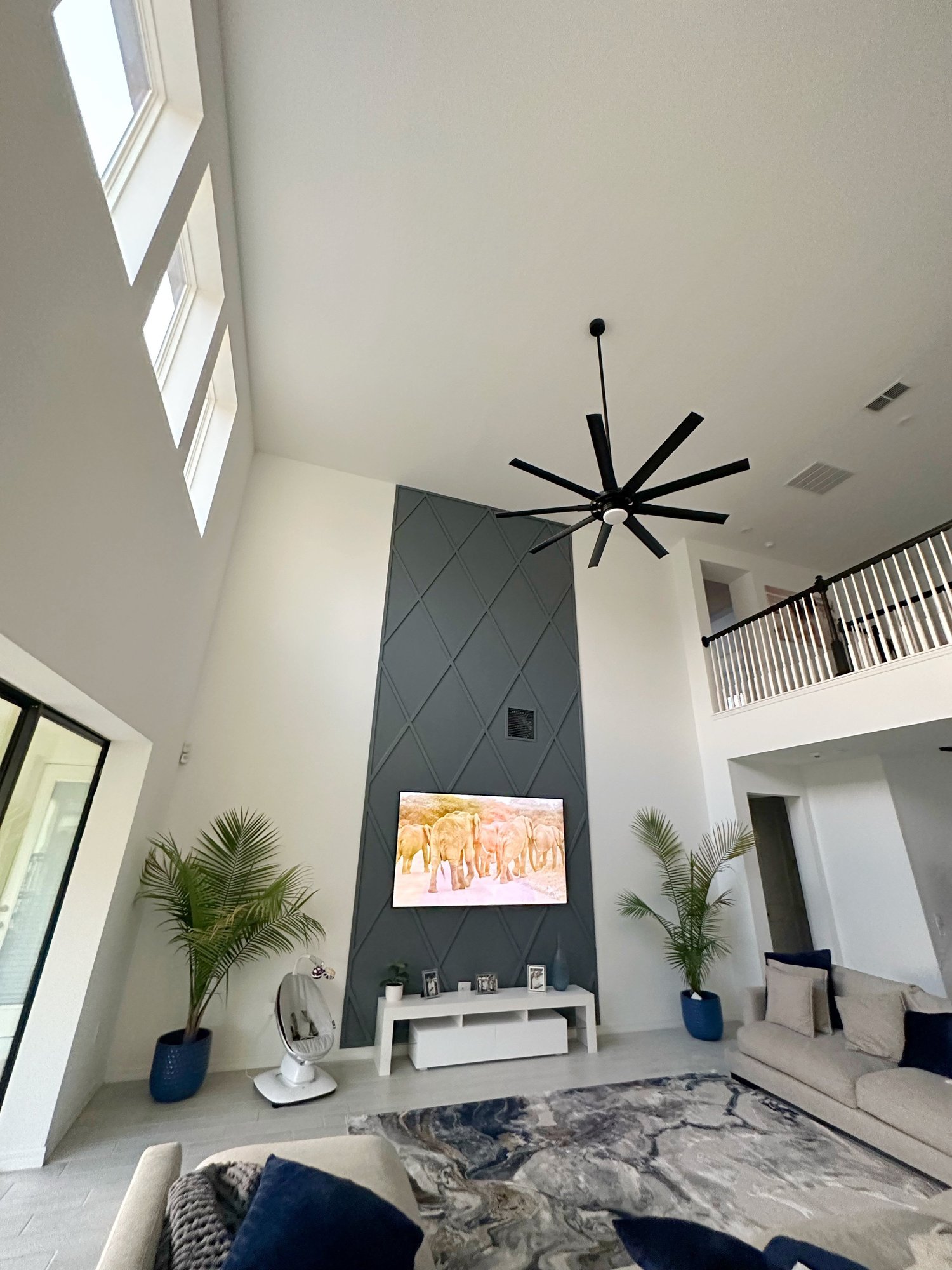 Modern living room with high ceiling, black ceiling fan, TV displaying elephants, gray accent wall, white entertainment unit, potted palms, beige sofa, and stone-patterned rug.