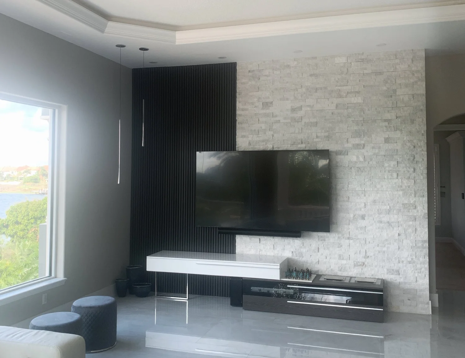 Modern living room with large window, black textured accent wall, flat-screen TV, white and black media console, light-colored stone accent wall, and round ottomans in front of a sofa.