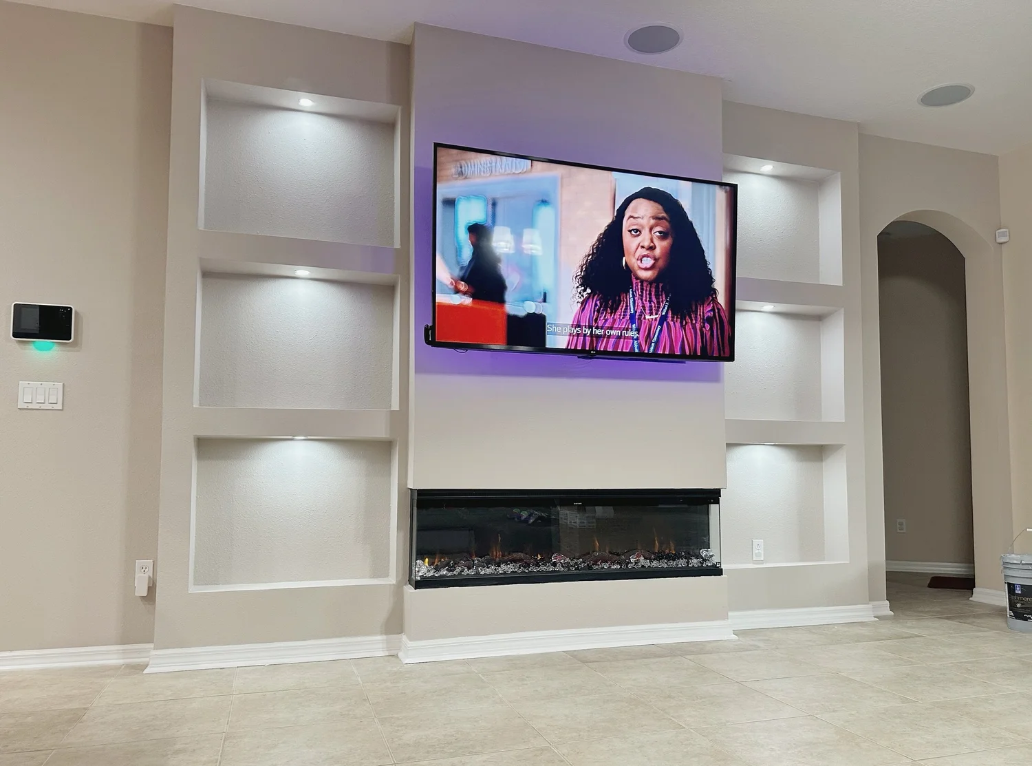 Living room with a wall-mounted TV showing a woman speaking, built-in shelving, a linear electric fireplace below the TV, and beige tiled flooring.