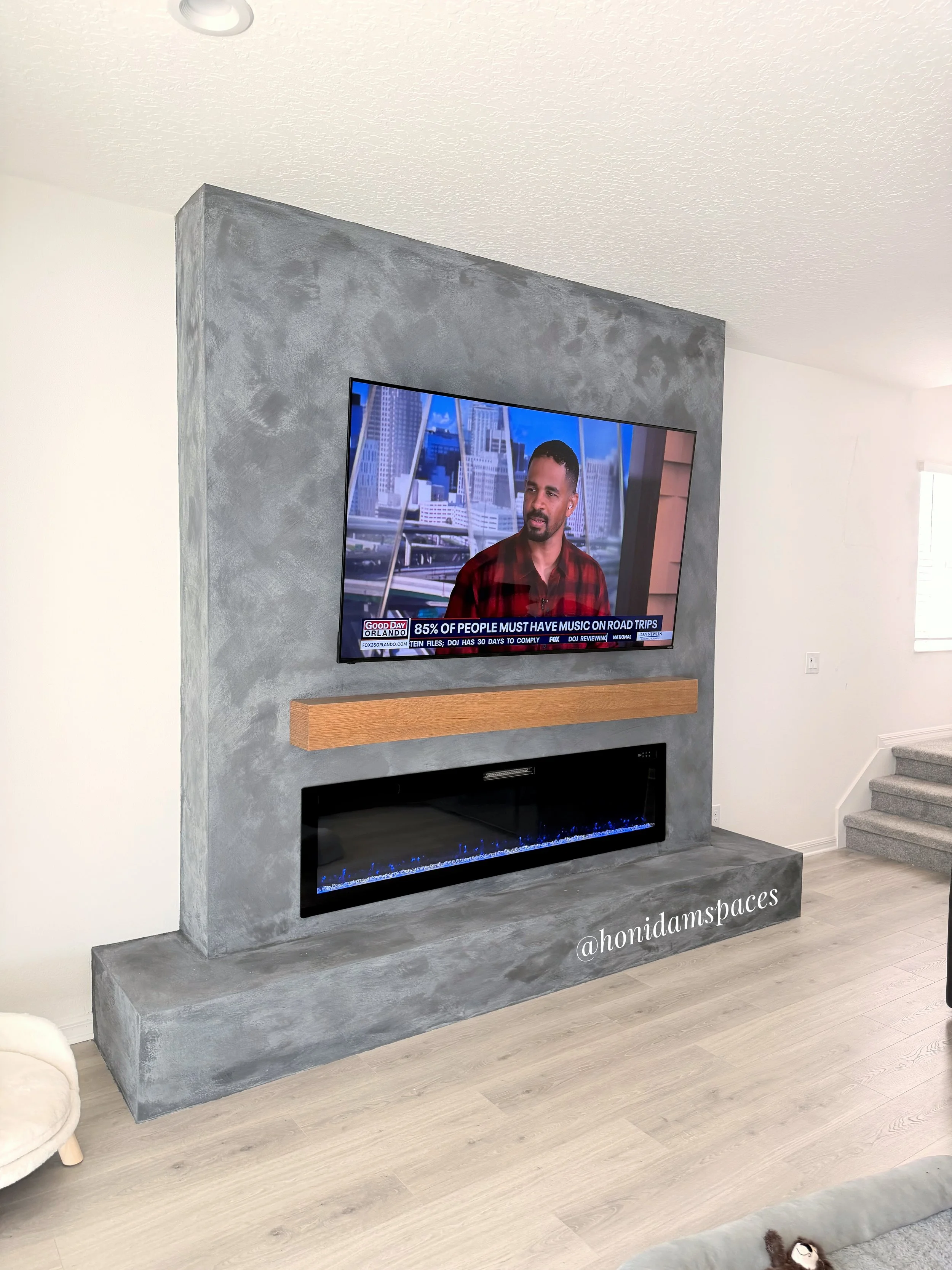 Living room with a modern gray textured fireplace wall, a flat-screen TV mounted above a wooden shelf, and a built-in electric fireplace with blue flames. The space features light wooden flooring, white walls, and a staircase with gray carpeting.
