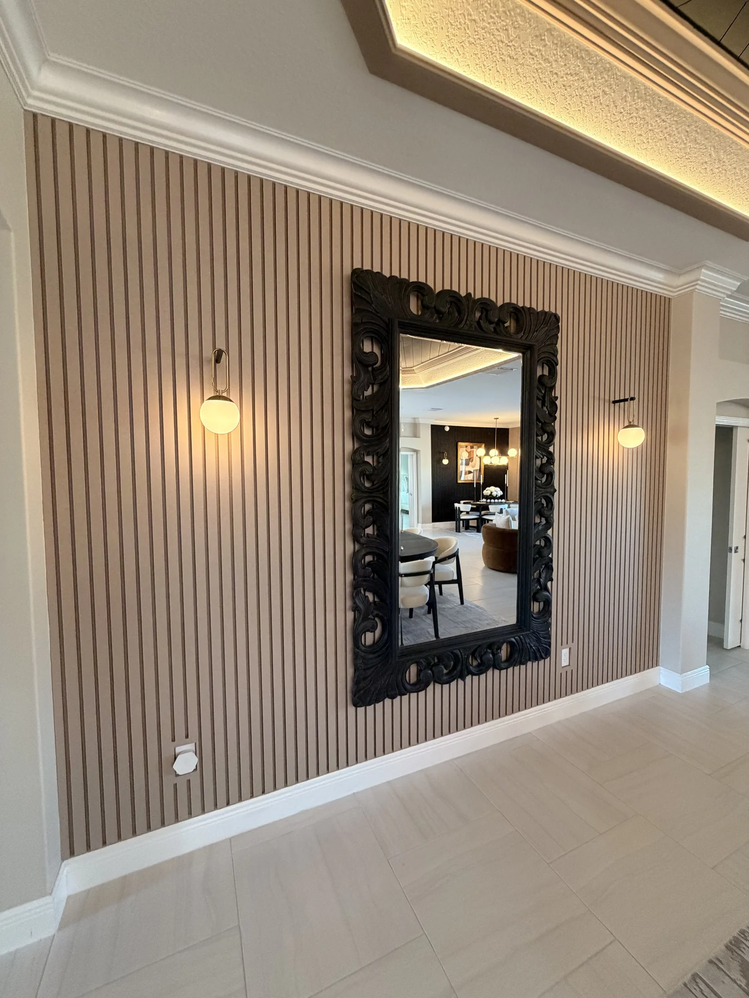 A large black ornate mirror hangs on a striped beige and brown wall in a modern living room, reflecting a dining area with a table, chairs, and a chandelier.