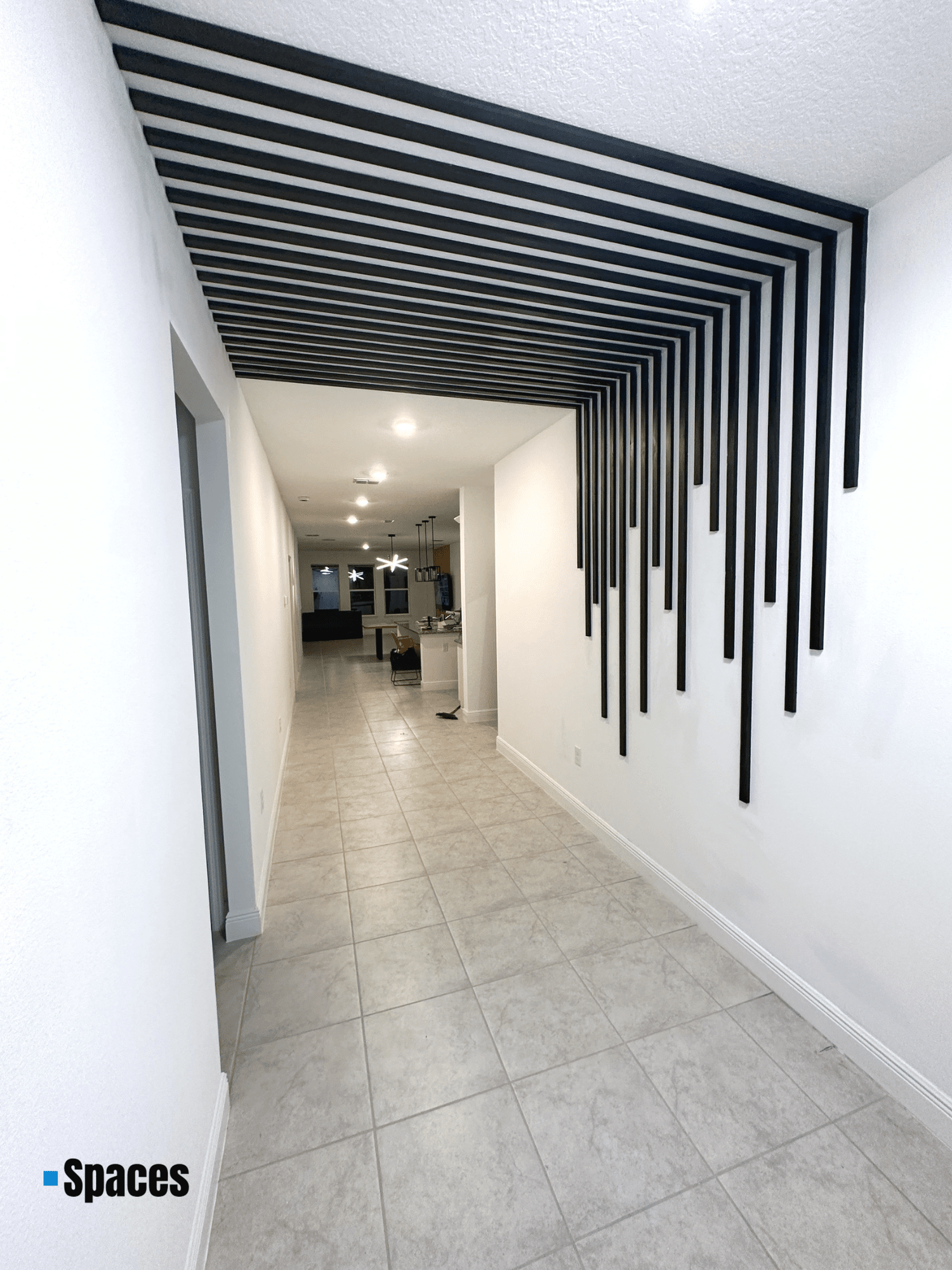 Empty hallway with white walls, beige tiled floor, black geometric wall art, and modern ceiling lights in a residential home.