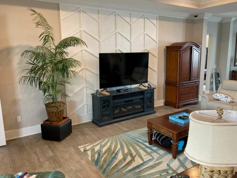 Living room with a large potted plant, a black TV stand with a flat-screen television, a wooden armoire, a beige armchair with a pillow, a wooden coffee table with a blue box, and a patterned area rug.