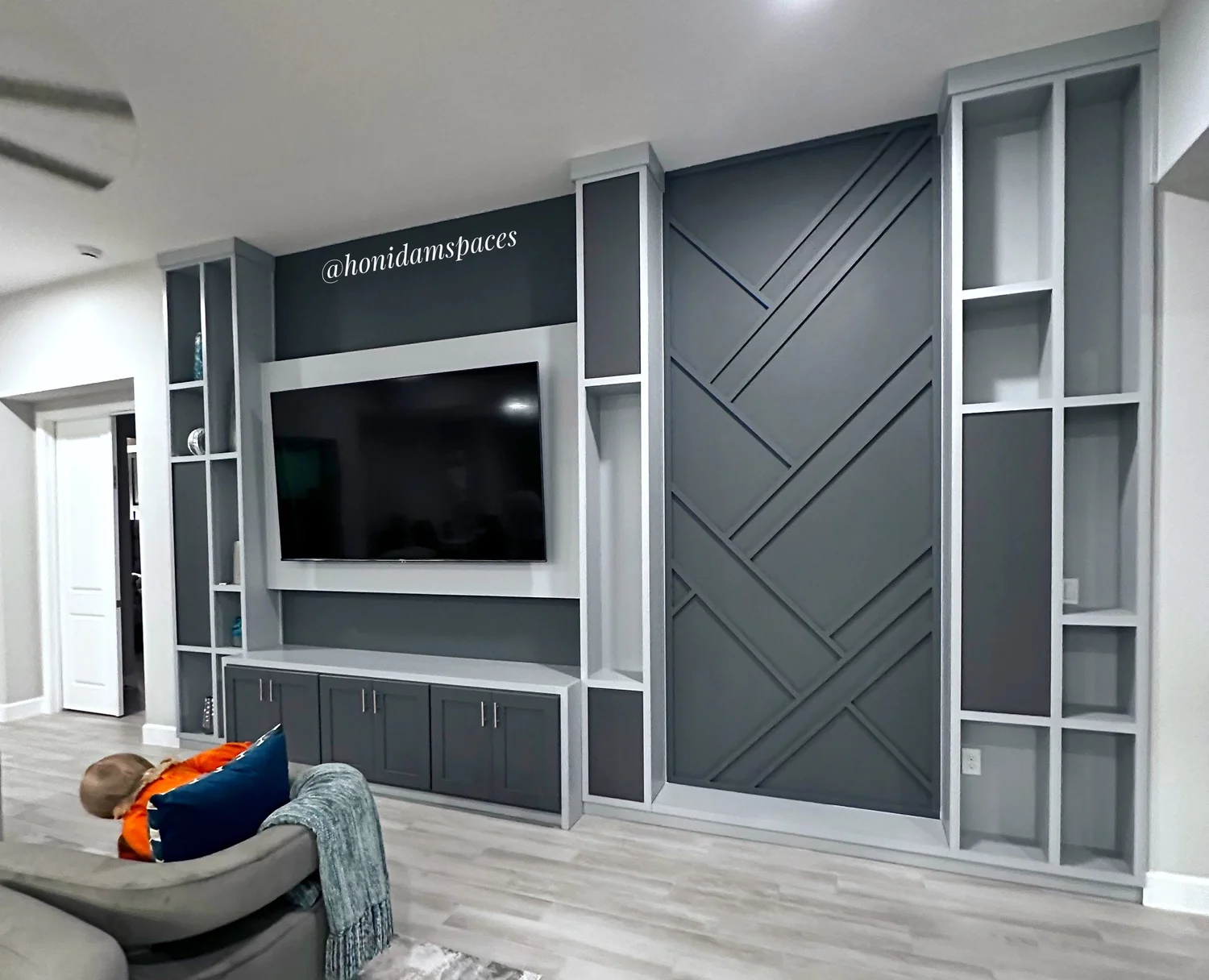 Living room with a large custom white and gray built-in entertainment center with shelves and drawers, next to a doorway, with a child lying on a sofa.