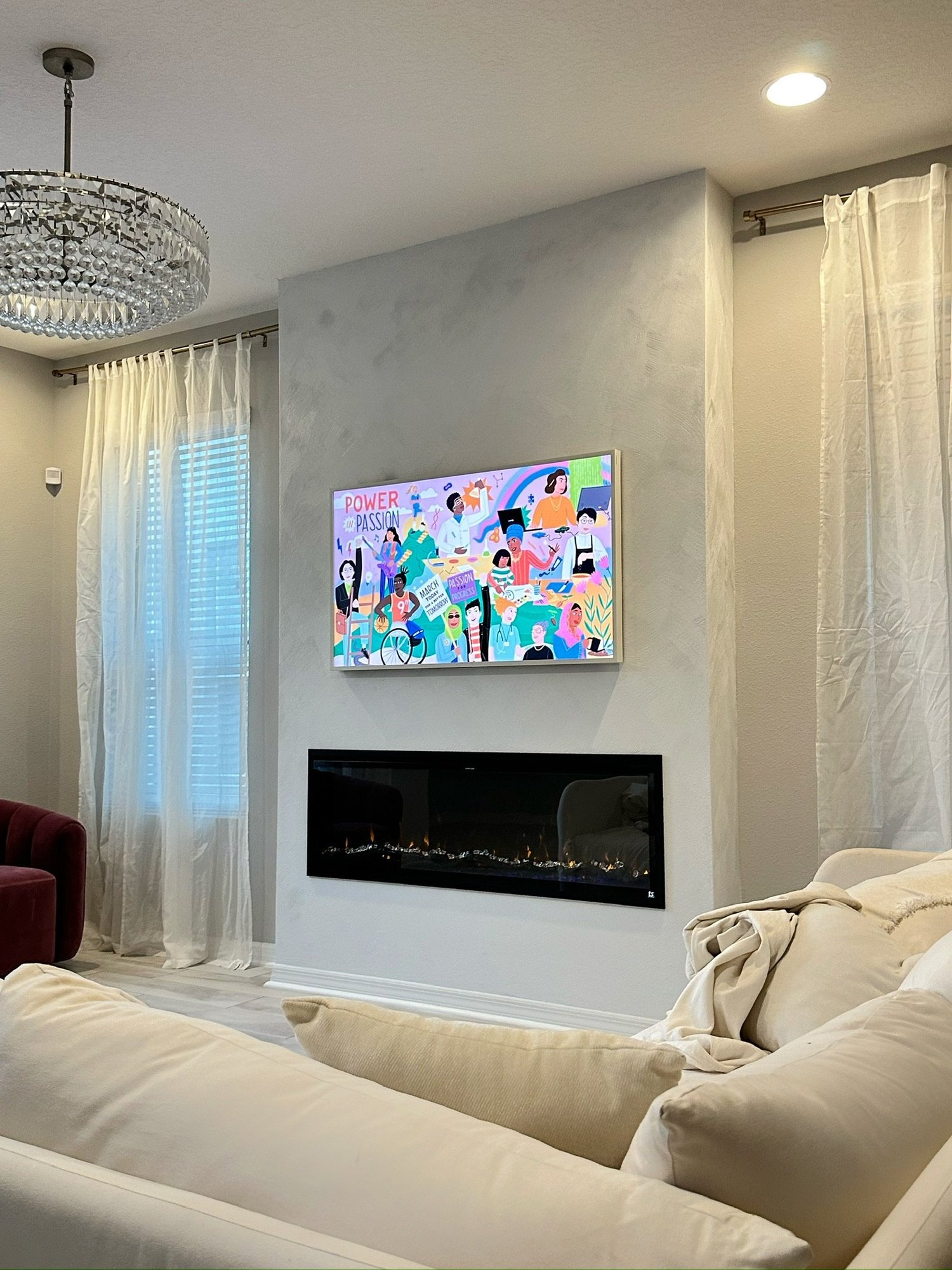 Living room with a wall-mounted TV showing an animated image of diverse women, a fireplace below, beige sofas, sheer curtains, a chandelier, and recessed ceiling lights.