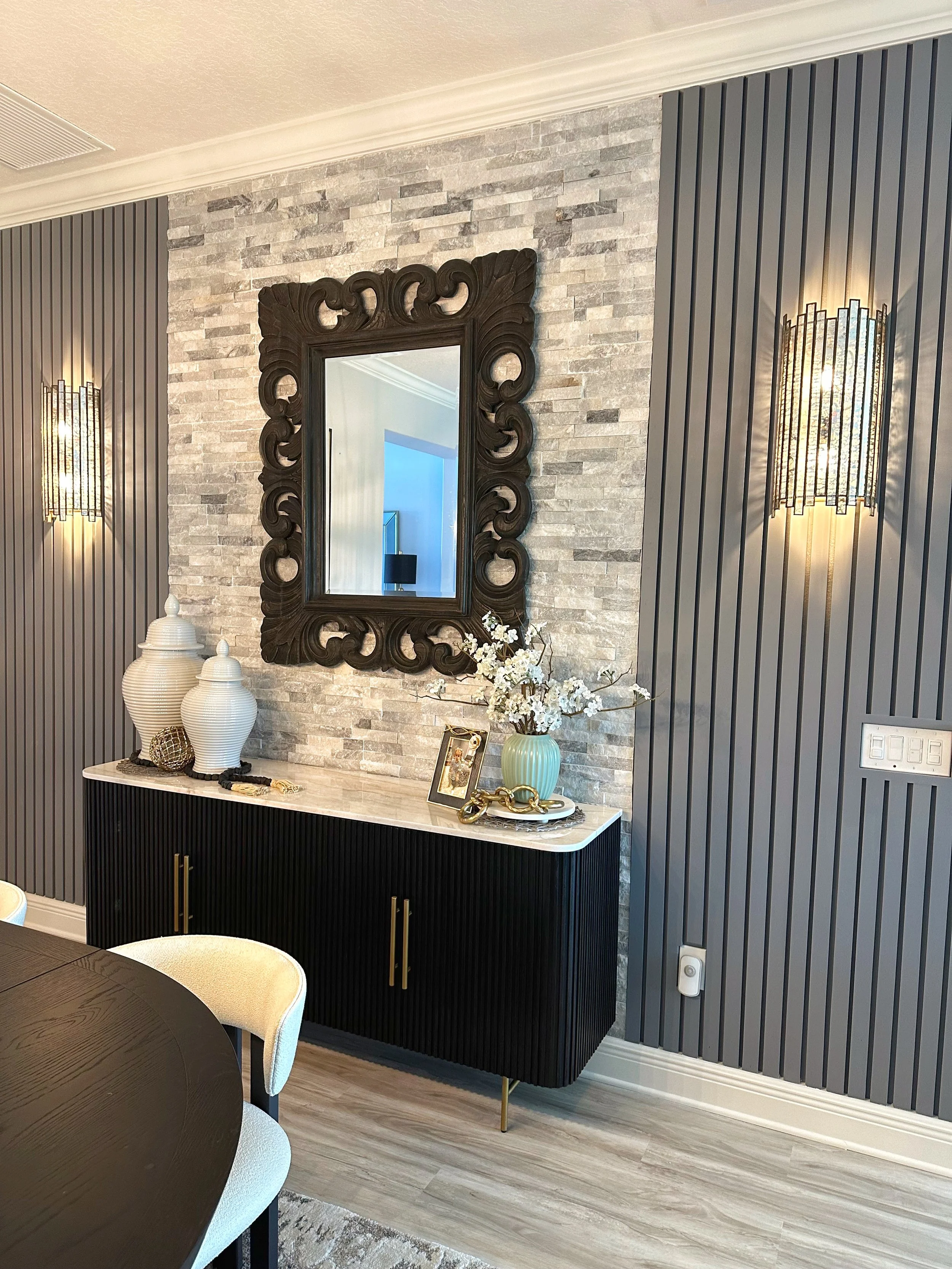 Decorative interior wall with dark wood framed mirror, striped wall panels, sconces, and a black sideboard with gold handles topped with vases, a framed photo, and floral arrangement.