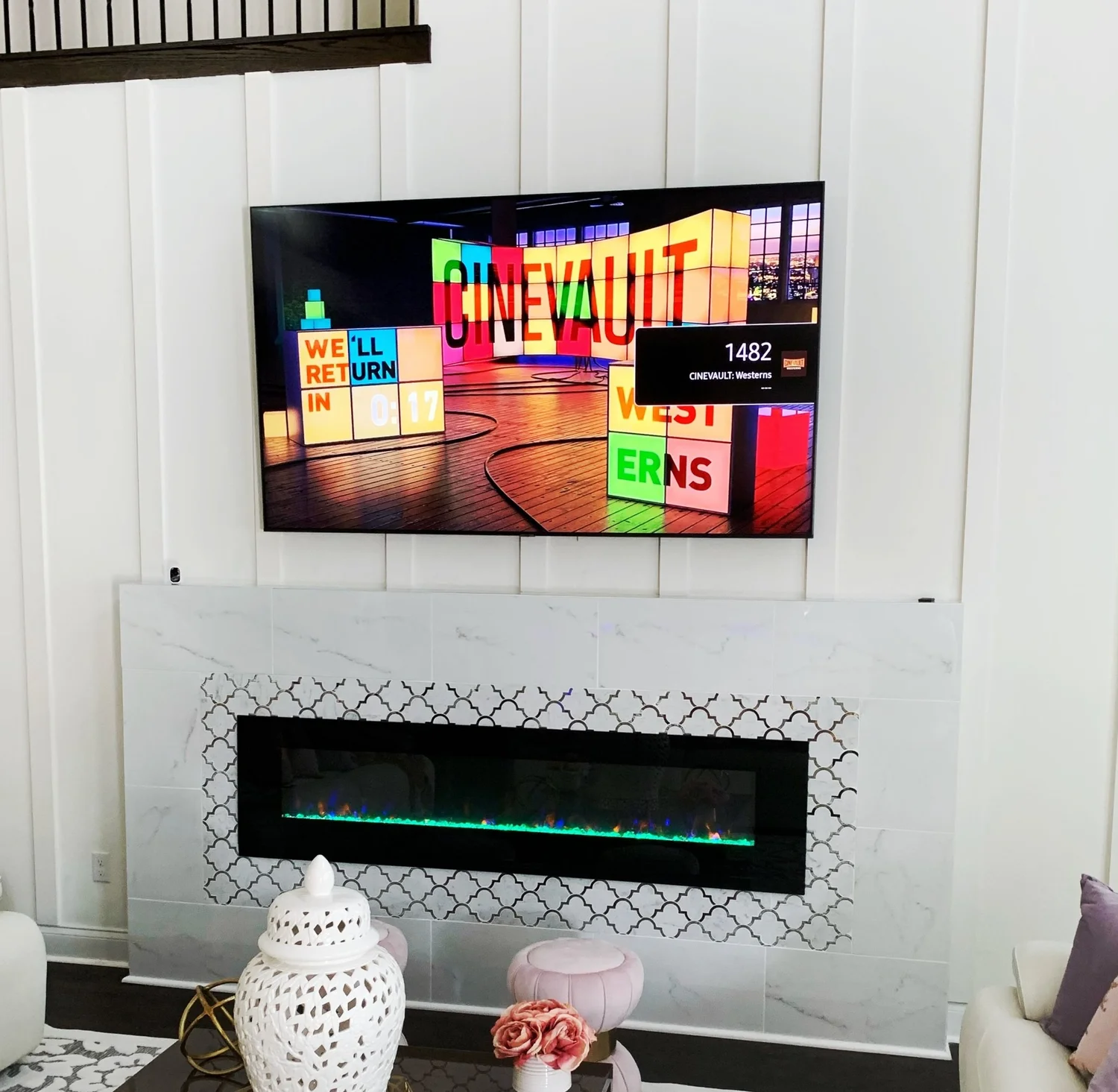 Living room with a mounted television displaying a colorful game show set and a fireplace below it with a decorative white urn and pink flowers on a table.