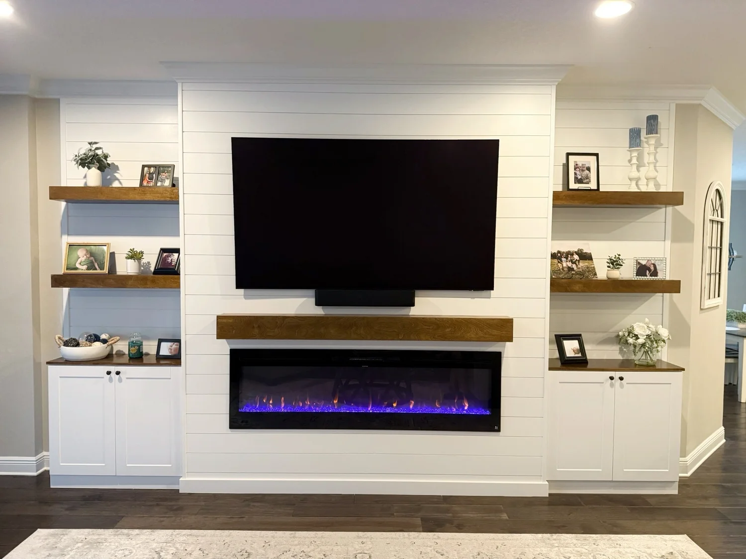 Living room with mounted flat-screen TV and electric fireplace on a white wall with wooden shelves and framed photos and decorative items.