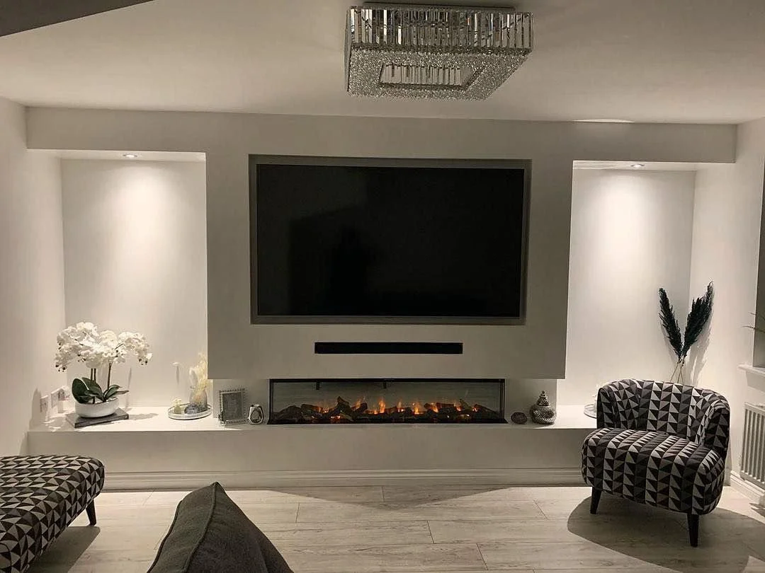 Living room with wall-mounted TV, electric fireplace, and decorative items. Two patterned armchairs are visible, one on each side of the fireplace, with a dark sofa partially in view. A modern chandelier hangs from the ceiling.