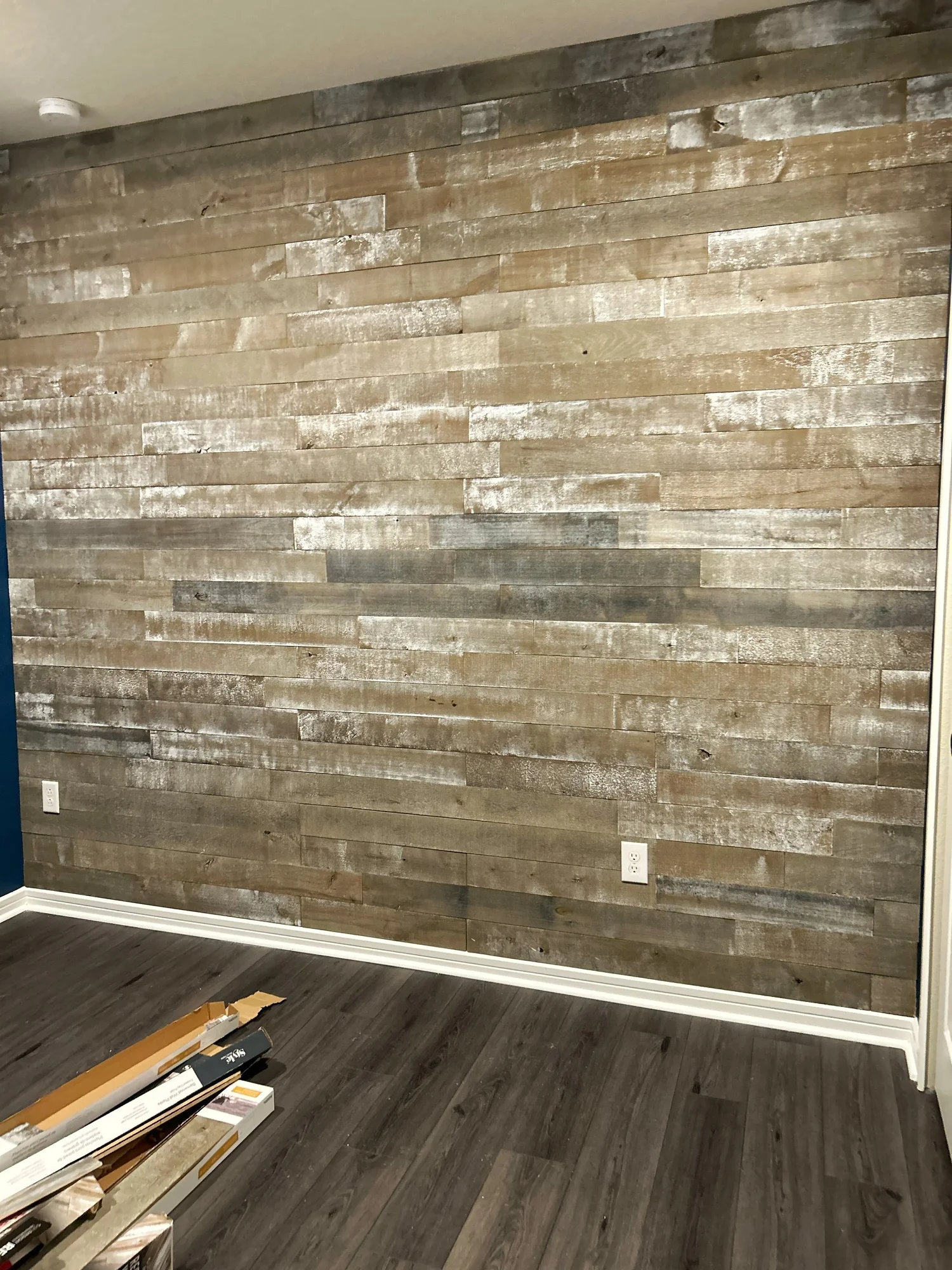 A room with a wooden accent wall, two electrical outlets, and a dark wood floor. There are some flooring samples or boxes on the floor in the corner.