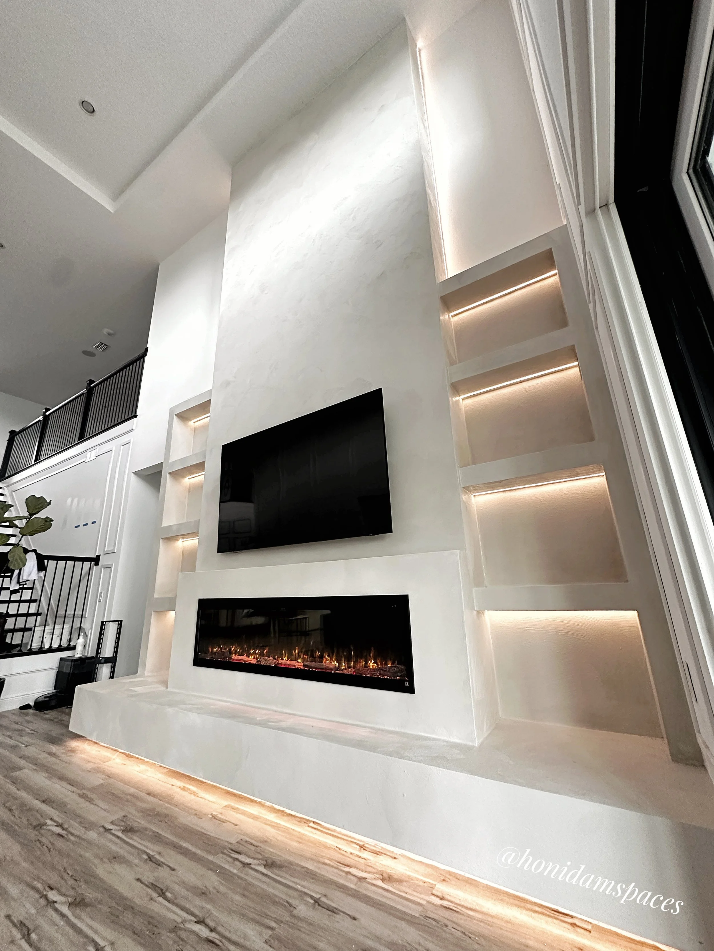 Modern living room with a large white built-in wall unit, a black wall-mounted television, and a fireplace below it. The wall unit features illuminated shelves and is illuminated at the base, with light-colored wood flooring.