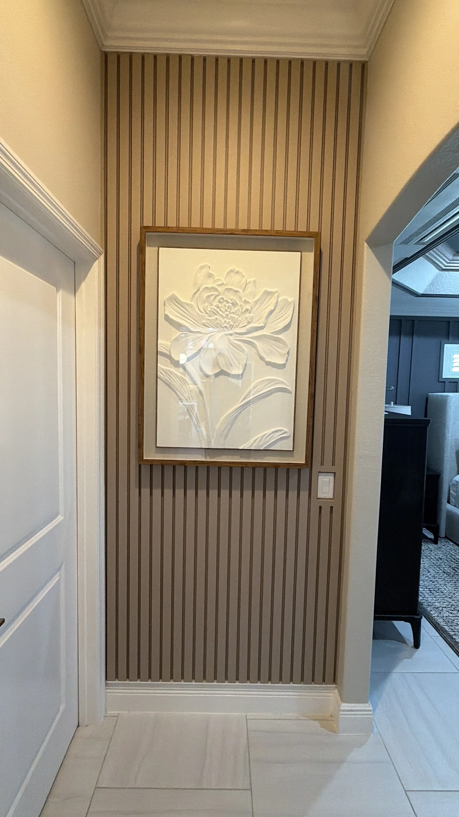 Hallway with striped wallpaper, a framed flower artwork, and a light switch, leading into a living room with a blue wall and white furniture.