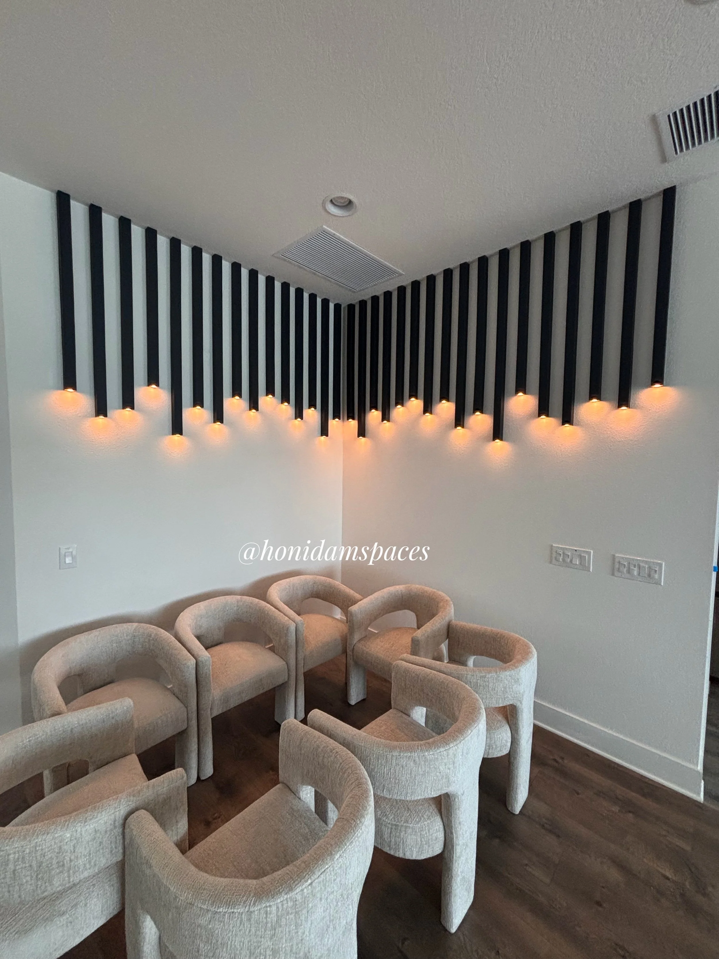 A room with a wall decorated with black vertical slats and orange accent lighting. There are beige upholstered chairs arranged in a circle, and wood flooring.