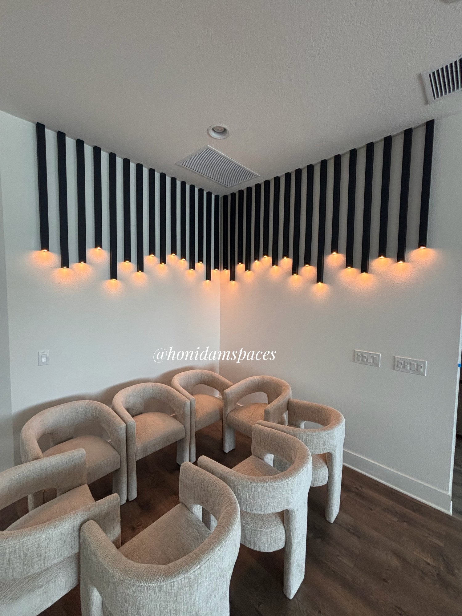 A room with a wall decorated with black vertical slats and orange accent lighting. There are beige upholstered chairs arranged in a circle, and wood flooring.