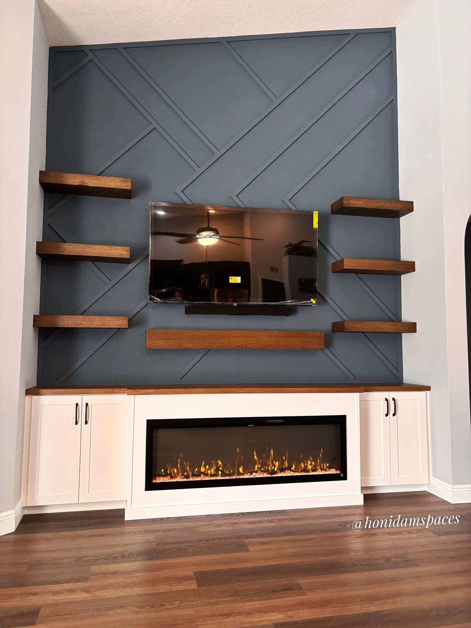 A living room corner with a modern electric fireplace with flames, white cabinetry, a mounted flat-screen TV, a decorative blue wall with diagonal wood paneling, and floating wooden shelves.