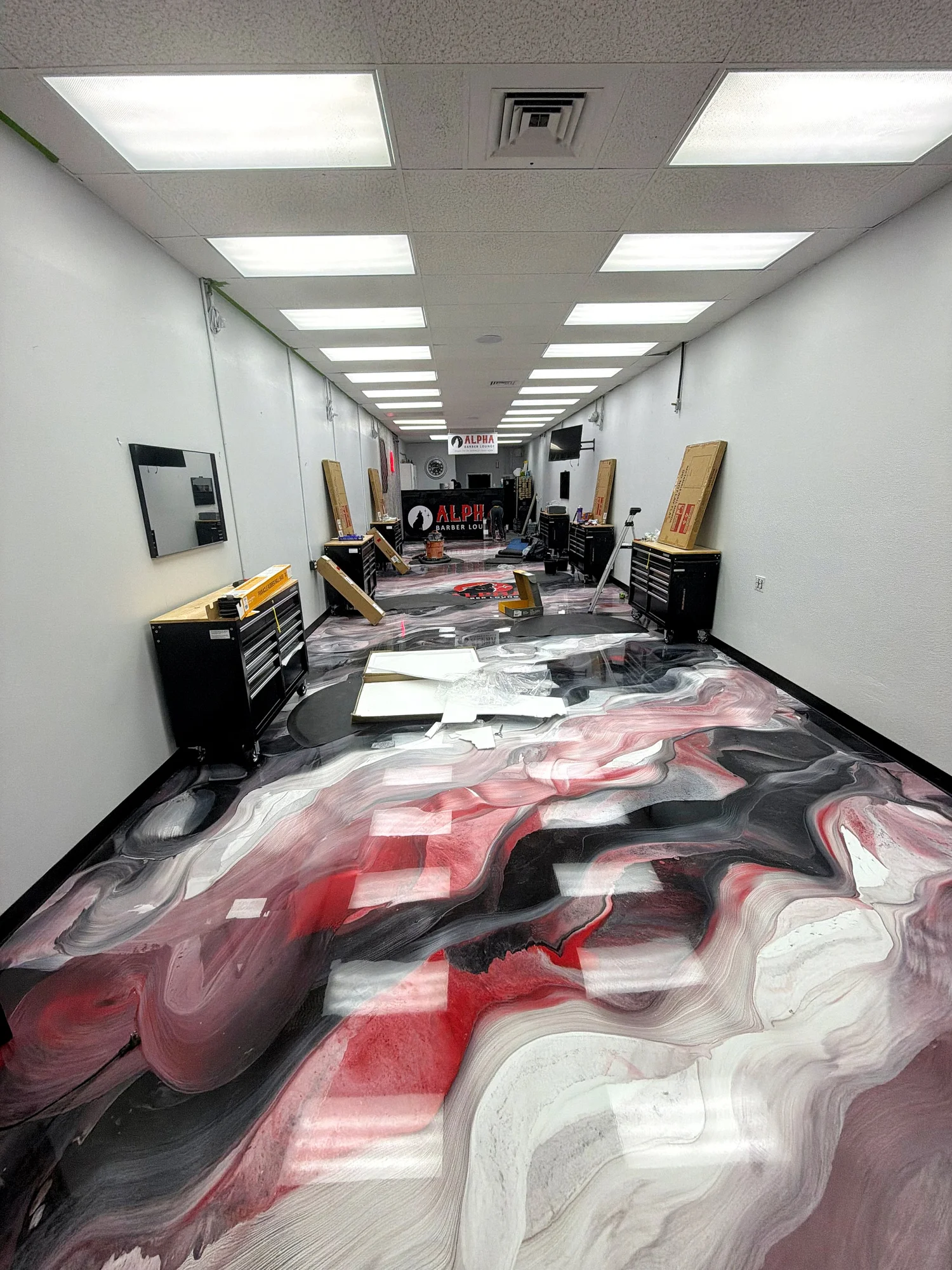 A long hallway with a marbled, red, white, black, and gray floor, lined with black tool chests and packaging materials, with a sign reading 'Alpha Barber Lounge' at the end.