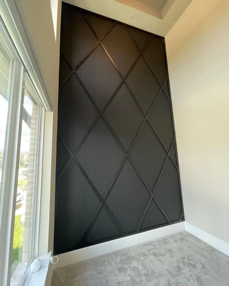 A black accent wall with a geometric diamond pattern in a room with beige walls, a large window, and light-colored carpet.