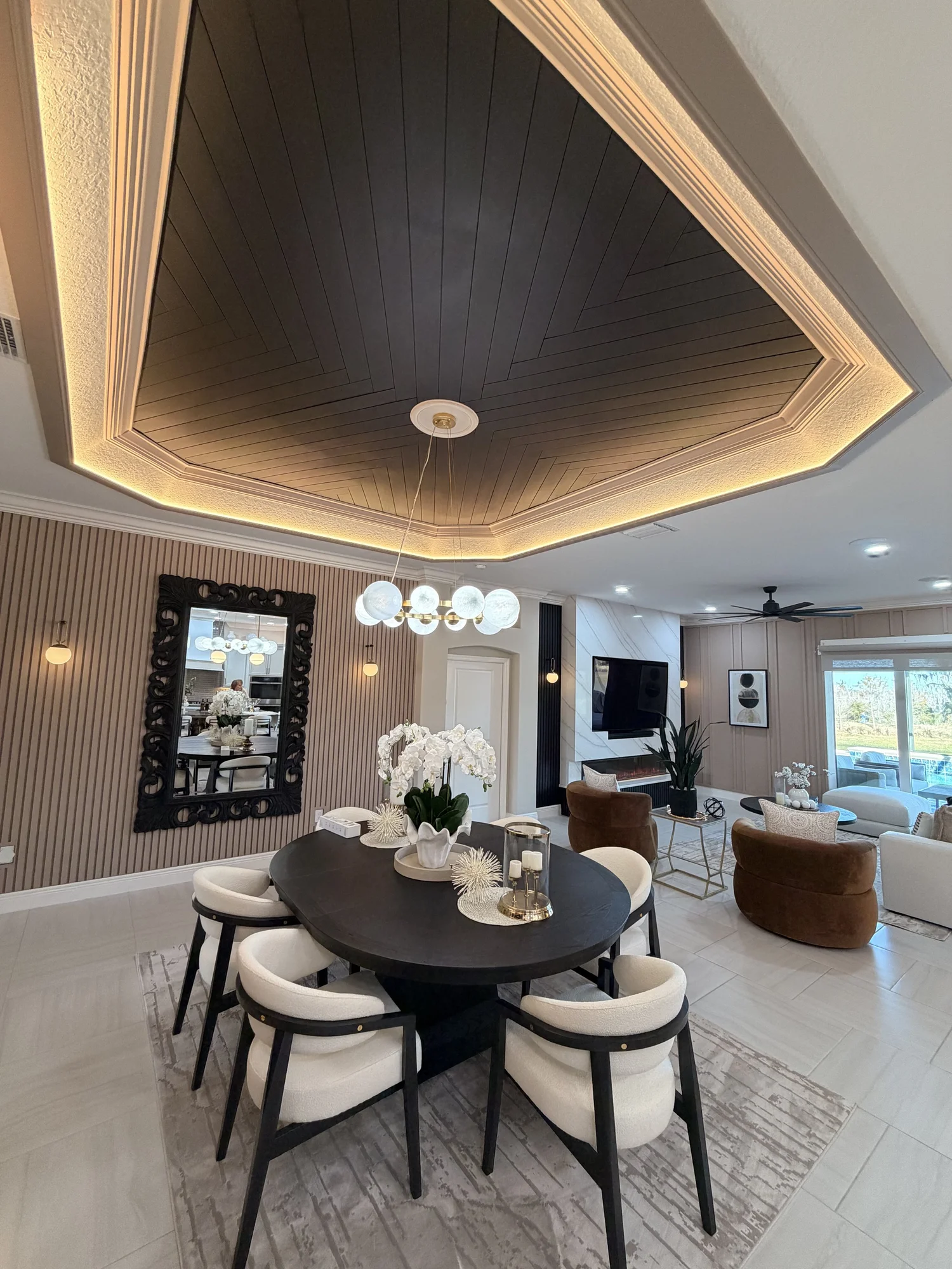 A living room with a dark round dining table surrounded by eight white chairs with black frames, a floral centerpiece, and a modern chandelier with several round globes. In the background, there is a wall mirror, wall sconces, a mounted TV, and a sea