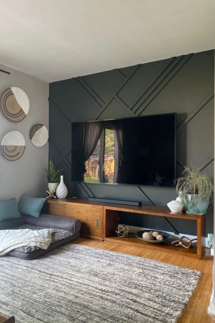 Living room with a wall-mounted TV, dark geometric-patterned wall behind it, two large potted plants, a wooden console, and wall art with round mirrors. A floor cushion with blankets and pillows is on a large striped rug on a wooden floor.